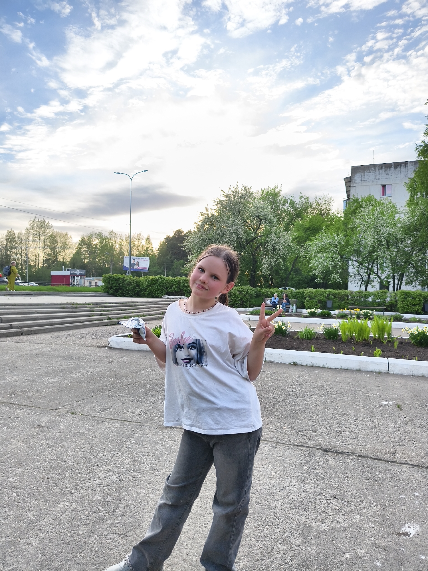 A young female is standing on a gray concrete surface, wearing a white t-shirt with a print, gray jeans, and gray shoes. She is holding an object in her left hand and making a peace sign with her right. Behind her, there are trees with white blossoms, green bushes, and a building in the background. A street lamp is visible on the left side of the frame. A concrete staircase is situated on the left side of the image and a group of people are sitting on a bench between the trees.