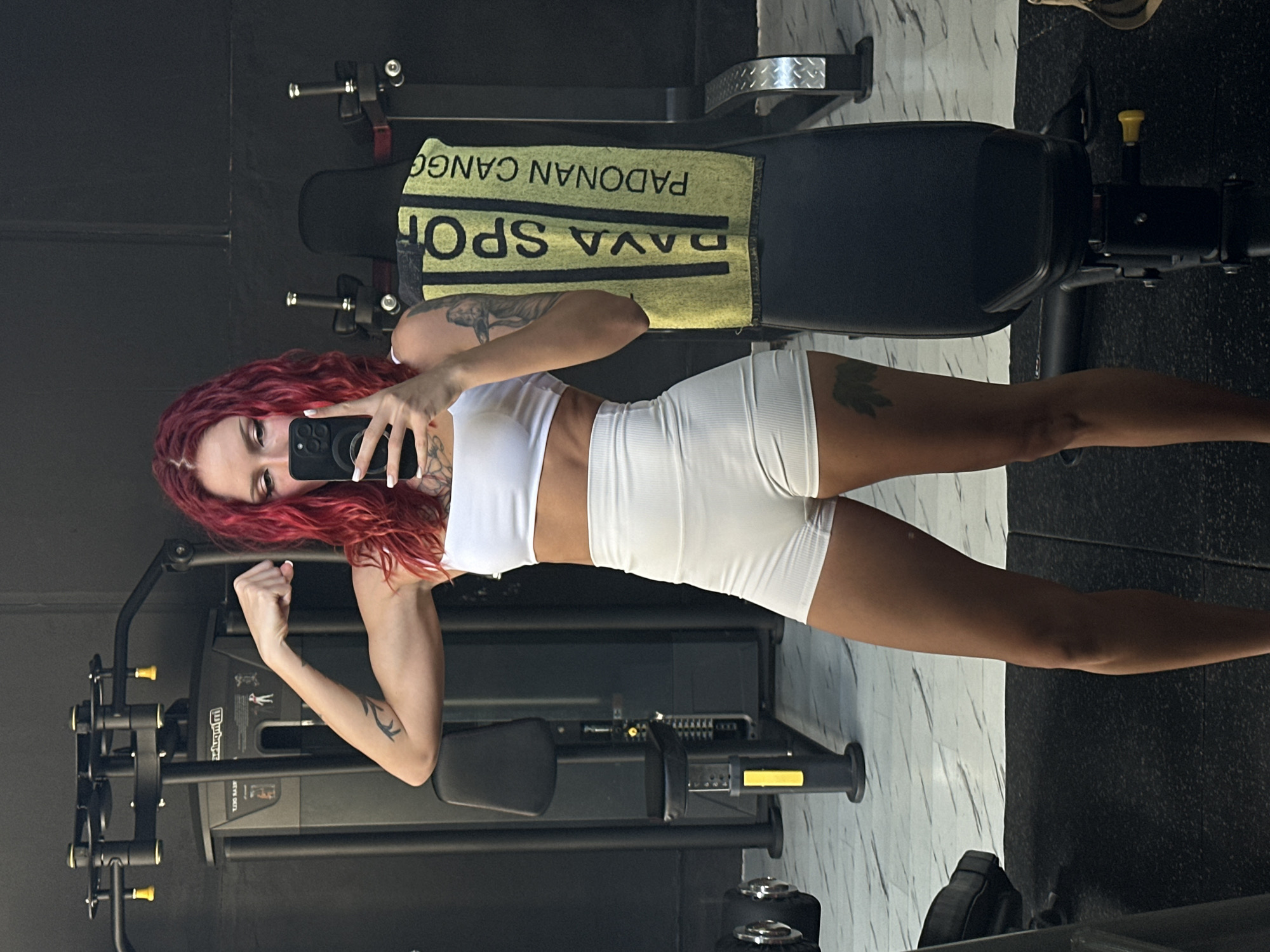 In the city of North Kuta, Indonesia, a woman with vibrant red, curly hair stands in a gym, posing for a selfie in a mirror. She is wearing a white athletic crop top and high-waisted white shorts. Her left arm is flexed, showcasing her bicep. She holds a black smartphone in her right hand, angled to capture her reflection. The background reveals various gym equipment, including weight machines and benches. A yellow towel with text in a foreign language hangs on one of the benches. The gym floor is a combination of marbled tiles and black rubber mats. The overall atmosphere is one of fitness and personal training.