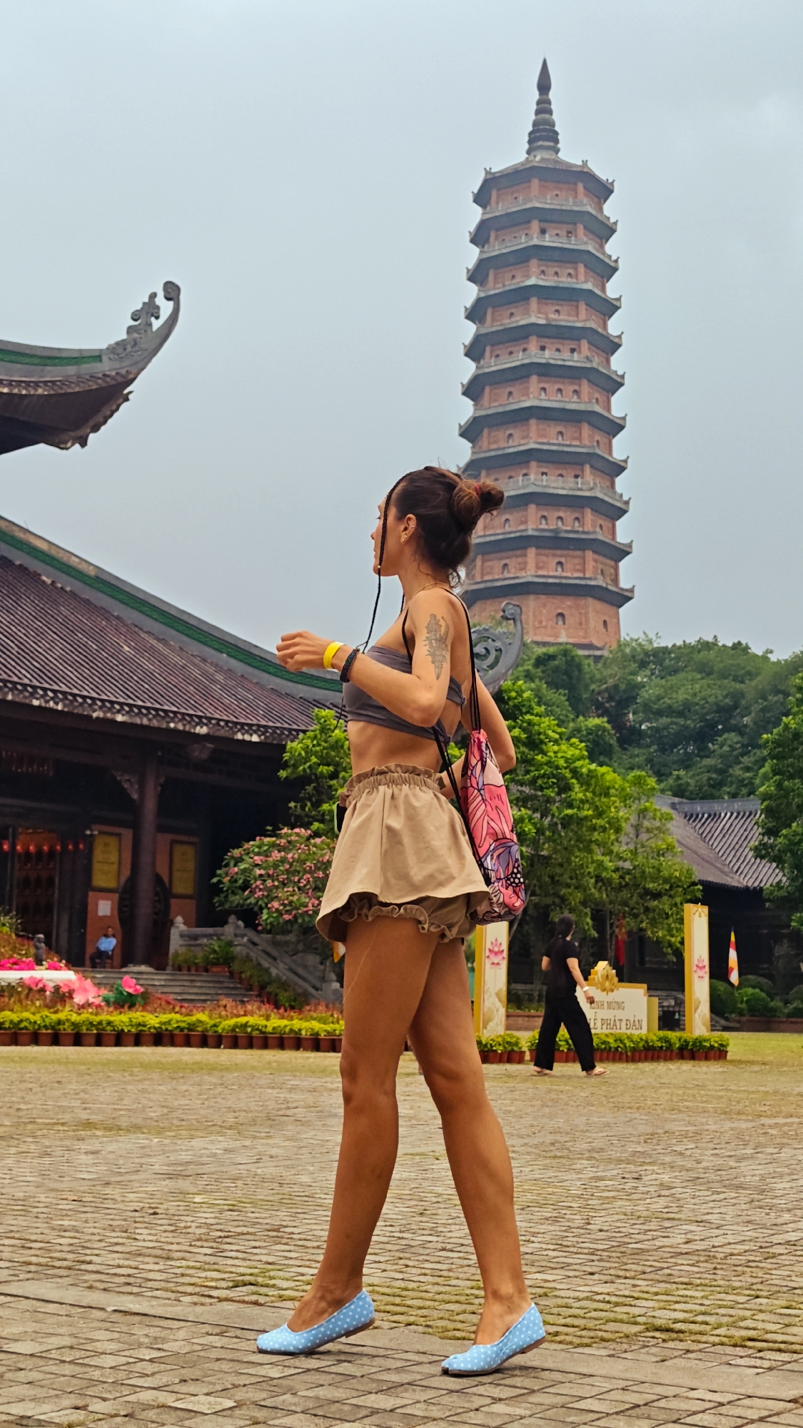 The image depicts a woman walking on a paved courtyard in front of a temple complex in Tây Hoa Lư Ward, Vietnam. She is wearing a gray crop top, a tan ruffled skirt, and blue polka-dot ballet flats. She has her hair in a bun with braids and is carrying a patterned drawstring bag over her right shoulder. A tall, multi-tiered brick pagoda is visible in the background, along with traditional temple architecture and lush greenery. In the middle ground, another person is walking away from the camera, and decorative potted plants line the pathways. The sky is overcast.