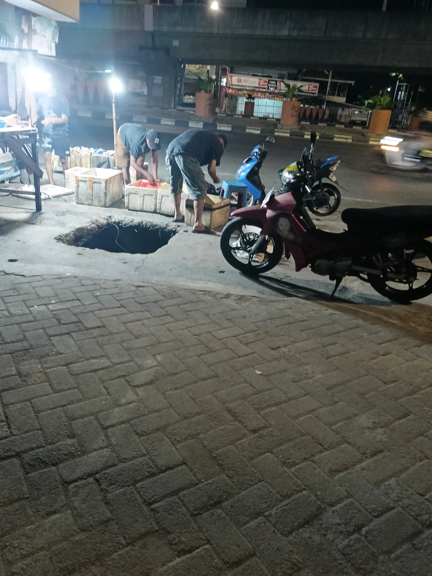 The image depicts an outdoor scene at night, possibly in Makassar, Indonesia, focusing on a street-level area near a road. In the foreground, a dark brick-paved walkway stretches diagonally across the frame. Adjacent to the walkway is a concrete surface with a large, dark, rectangular hole in it.
Several people are visible near the hole and a table on the left side of the image. One person, wearing a dark t-shirt and shorts, stands by the hole. Another individual, also in dark clothing and a baseball cap, is bent over a large rectangular container, seemingly engaged in some task. A third person is partially visible in the background, standing by the table.
Several large, white rectangular containers are placed around the hole, some of which appear to hold goods. A bright light source, possibly a streetlamp, illuminates the immediate area.
In the mid-ground, two motorcycles are parked. A blue motorcycle is situated behind a dark red motorcycle, which is closer to the foreground. The red motorcycle has a sticker on its side that appears to depict a child's face. The street beyond the motorcycles is visible, with blurred lights indicating passing vehicles.
In the background, a bridge or overpass structure is present, with a building or storefront visible underneath it. The environment suggests a commercial or public area operating at night, with elements of urban infrastructure and street vending or activity.