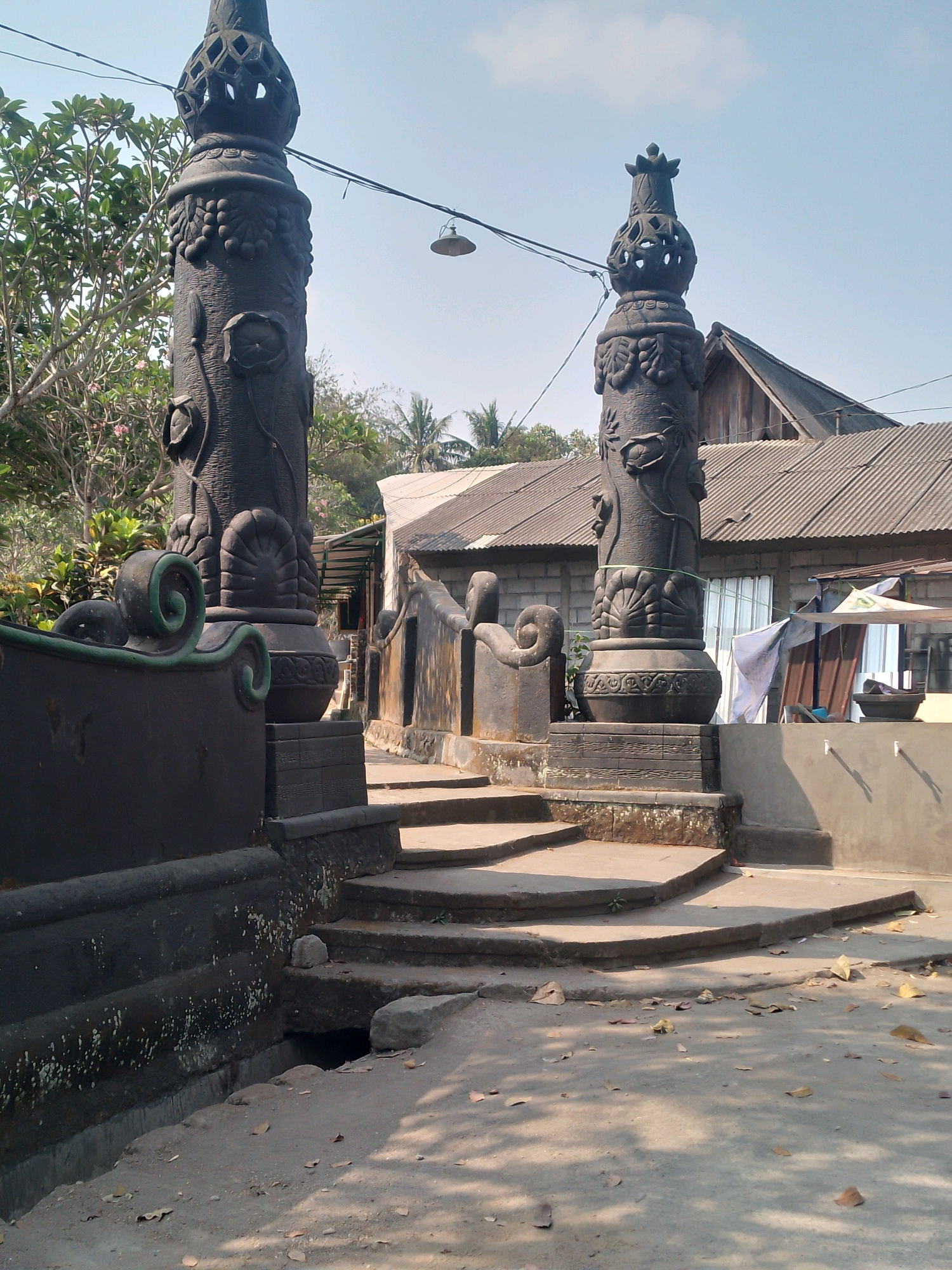 In Gunungpring, Indonesia, an outdoor scene features two tall, intricately carved stone pillars, possibly decorative elements of an entrance or monument. The pillars are dark in color and adorned with relief carvings of what appear to be grapes, lotus flowers, and other botanical motifs. The pillar on the left is closer to the viewer and is partially obscured by a dark, curved wall or railing with green decorative trim. Steps made of concrete lead upwards between the pillars.
In the background, a building with a corrugated metal roof is visible, with various structures and tarpaulins suggesting a utilitarian or storage area. Trees with lush green foliage and some blooming pink flowers are present to the left of the pillars, and palm trees are visible further in the background. The sky is clear and blue. The ground in the foreground is made of concrete and dirt, scattered with dry leaves. A small, dark opening in the ground is visible near the base of the steps, possibly a drain or opening to an underground space. There are no people present in the image. The overall impression is one of an outdoor cultural or historical site, possibly with elements of religious significance.