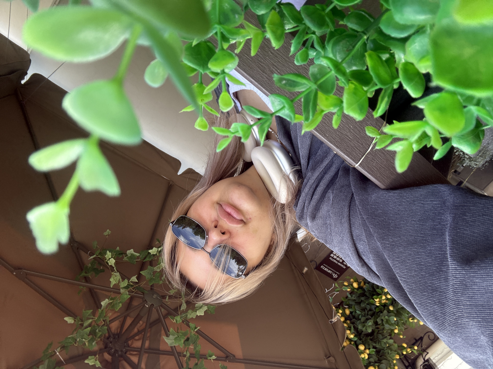 A young woman with blonde hair and sunglasses poses for a selfie outdoors. She is wearing headphones around her neck and a gray hooded sweatshirt. The background features a brown umbrella, artificial greenery, and a string of yellow lights. A sign with Russian text is visible on a building in the background. The lighting suggests it is daytime. The location is the city of Birobidzhan, Russia.