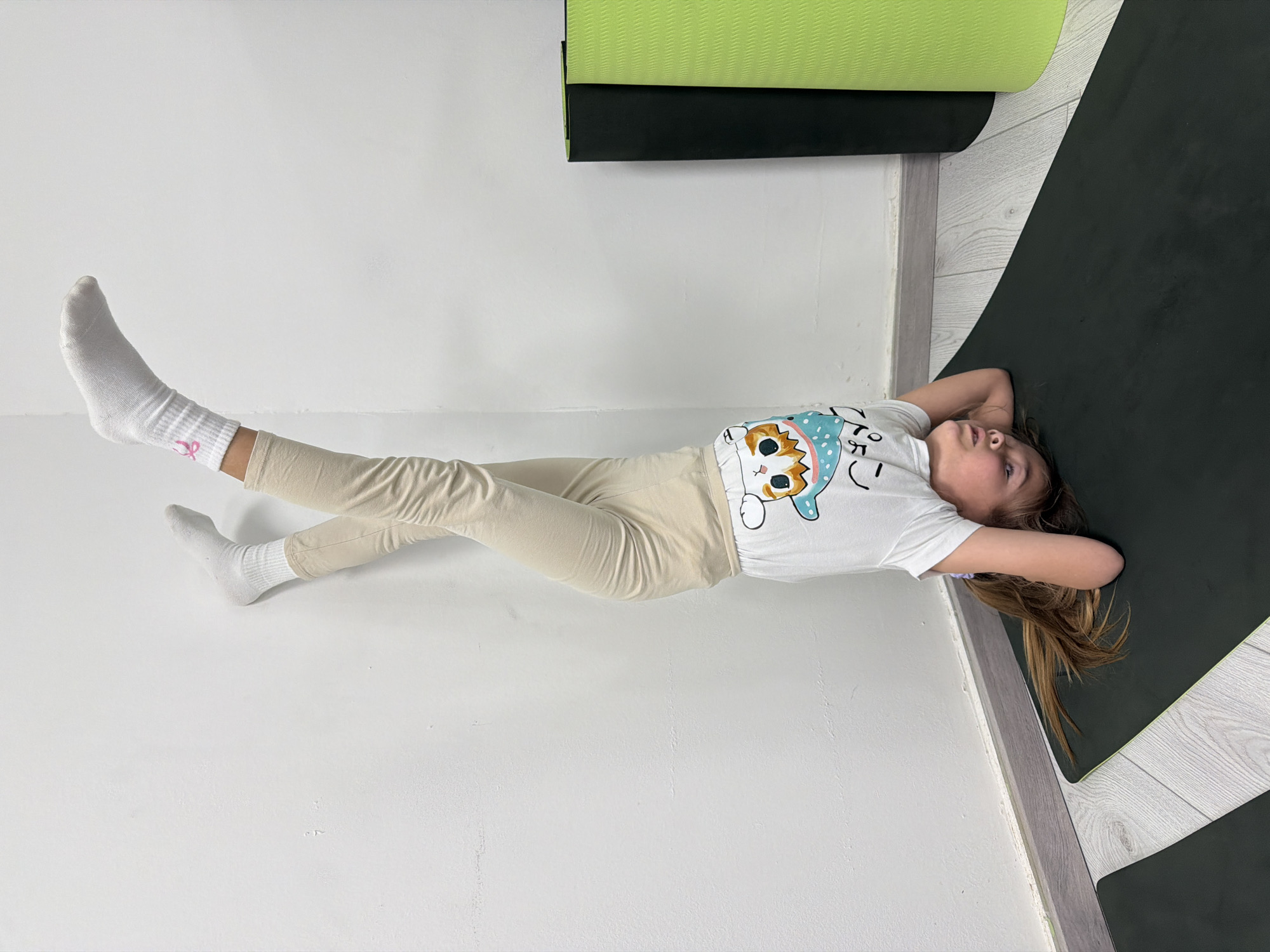 A young girl lies on a black yoga mat, her head near a wall and her body extending diagonally across the frame. She wears a white t-shirt with a cartoon cat design, beige pants, and white socks with pink accents. Her hands are behind her head. A rolled green yoga mat is visible near the top of the image. The background includes a white wall and a section of light wood-patterned flooring. The image does not clearly indicate the location is Budva, Montenegro.