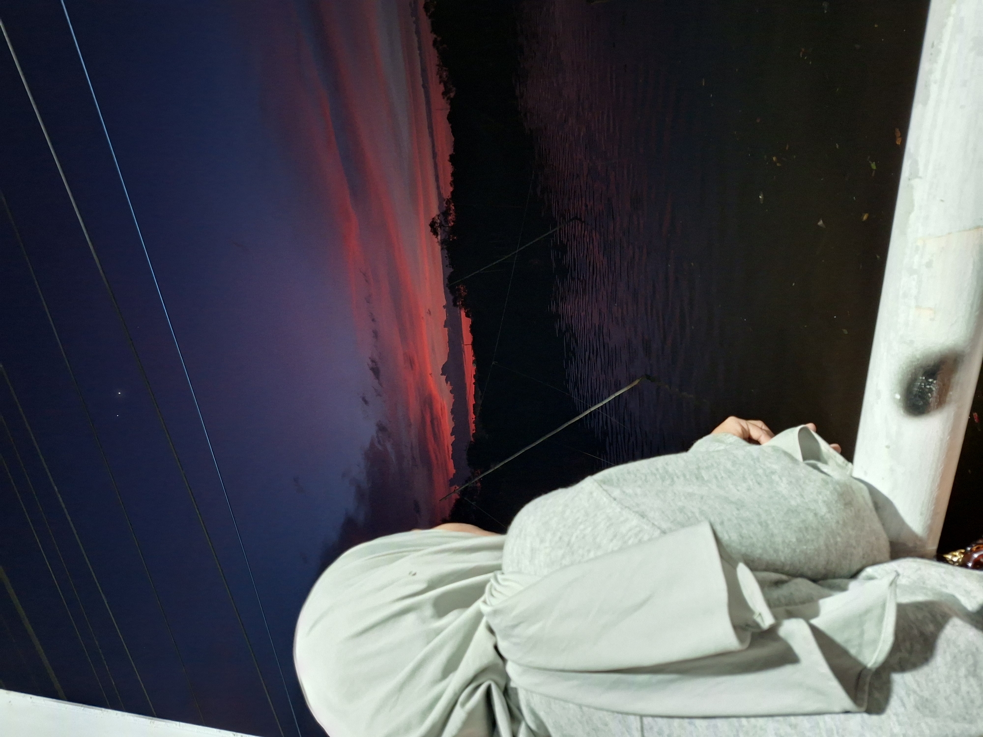An individual wearing a light grey hijab and hoodie stands by a railing, looking out at a body of water during sunset. The sky is a deep blue transitioning to vibrant shades of pink and orange from the setting sun, with scattered clouds. The water reflects the colors of the sky and appears dark. Silhouetted trees line the opposite bank of the water. Several thin, vertical poles with wires stretched between them are visible in the water. The railing in the foreground is white and shows signs of wear. This scene is likely located near the Krueng Cunda River in Lhokseumawe, Indonesia, a common spot for observing sunsets.
