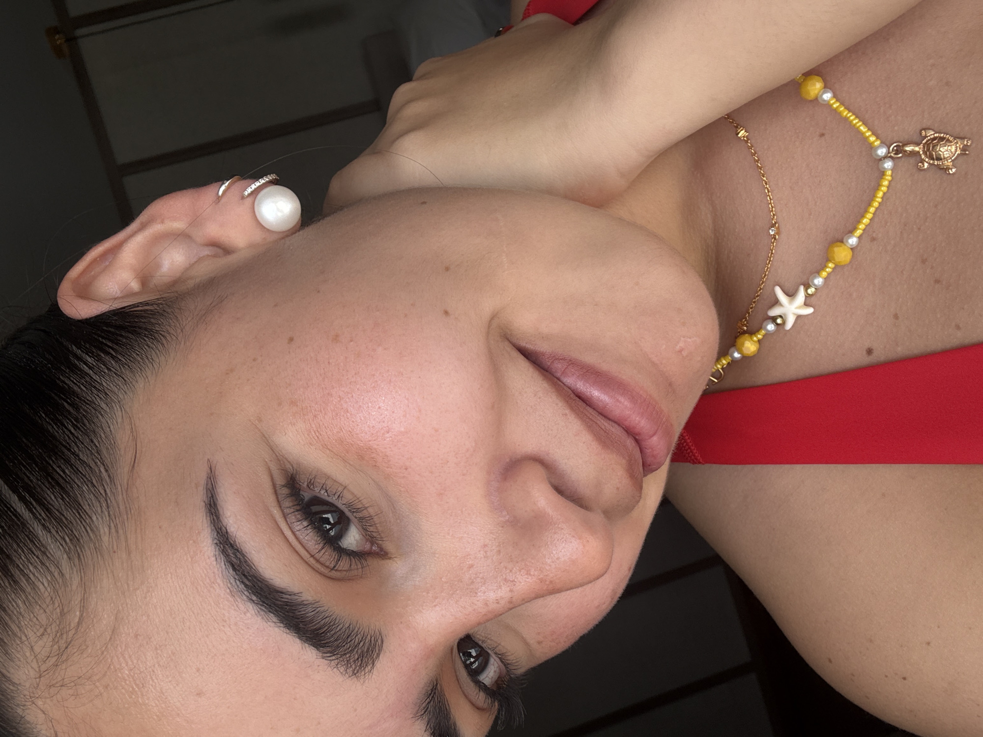 A close-up, tilted portrait features a woman with dark hair and fair skin. She has dark eyes and eyebrows, and light pink lipstick. Her left ear is visible, adorned with a pearl earring and a gold hoop. A yellow and white beaded necklace with a gold turtle pendant and a starfish charm rests on her chest, which is covered by a red garment. The background is dark and indistinct. The location is not discernible from the image.