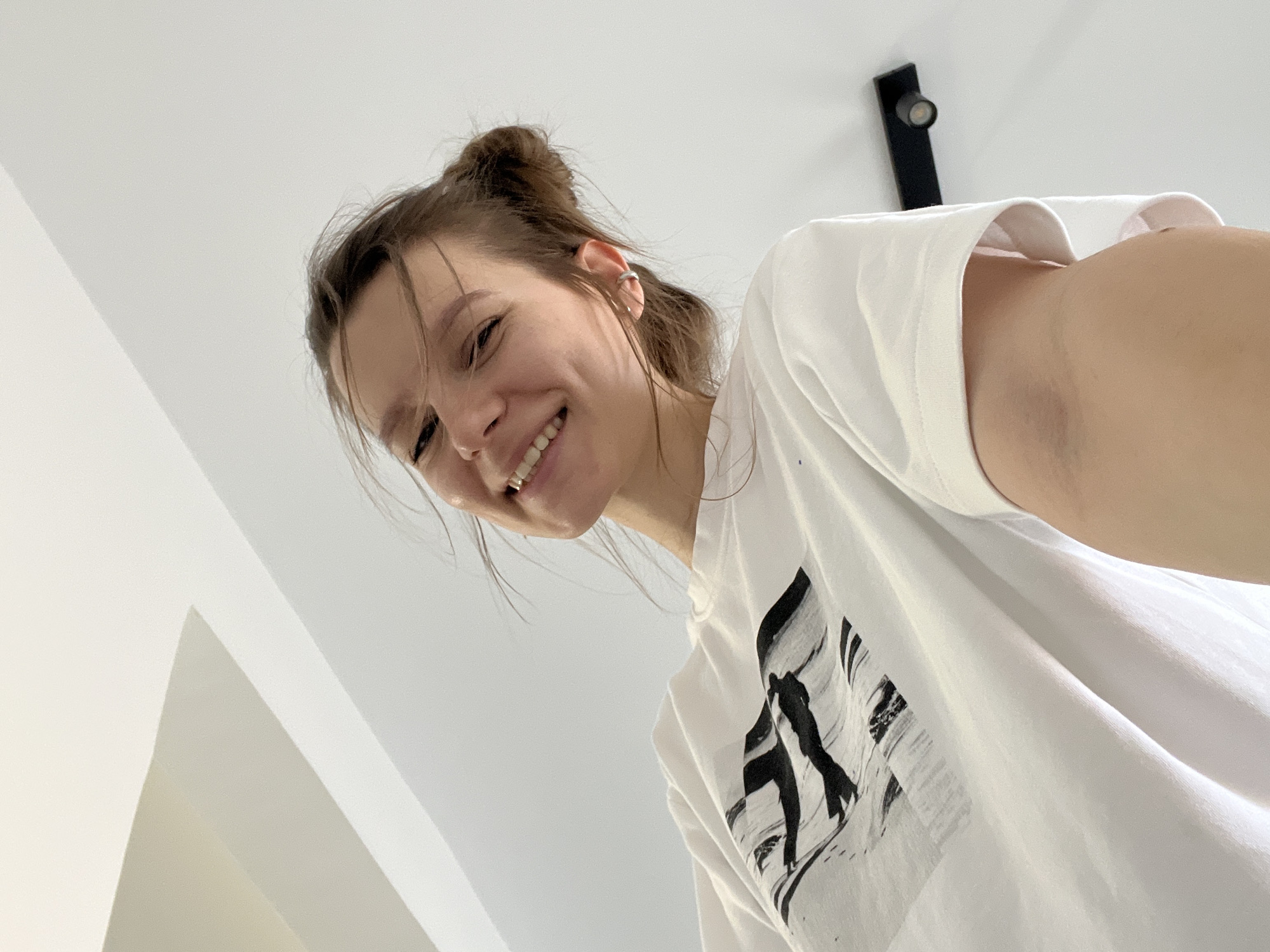 A low-angle, point-of-view shot shows a young woman with her hair pulled up in a messy bun, smiling broadly. She is wearing a white t-shirt with a black graphic design featuring abstract shapes and silhouetted figures. The woman is looking down towards the camera, her head tilted slightly. Her right ear, adorned with a small hoop earring, is visible. The background is a plain, bright white ceiling with a minimalist black light fixture mounted on it. The lighting suggests it is daytime, likely indoors. The overall mood is cheerful and informal. The image appears to be a selfie taken from a low perspective, perhaps while lying down or standing over something. The setting is clean and modern. There is no visible text in the image. The context of Limassol, Cyprus is not directly evident from the image itself but provides a geographical setting.