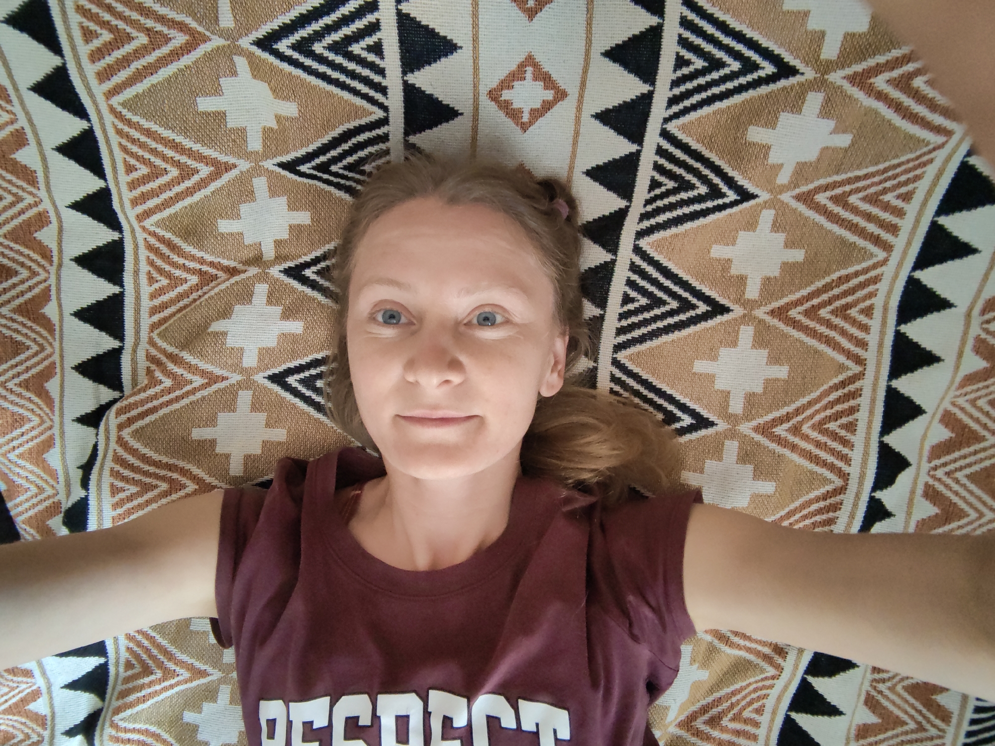 A young woman with blonde hair, tied in two pigtails with pink elastics, lies on her back on a patterned blanket. She is wearing a maroon t-shirt with the word "RESPECT" printed in white letters. The blanket features a geometric pattern with triangles and diamond shapes in black, white, and brown. Her arms are outstretched to her sides, and she is looking up at the camera. The scene appears to be indoors, on a bed or couch, in Зуйское сельское поселение, Ukraine.