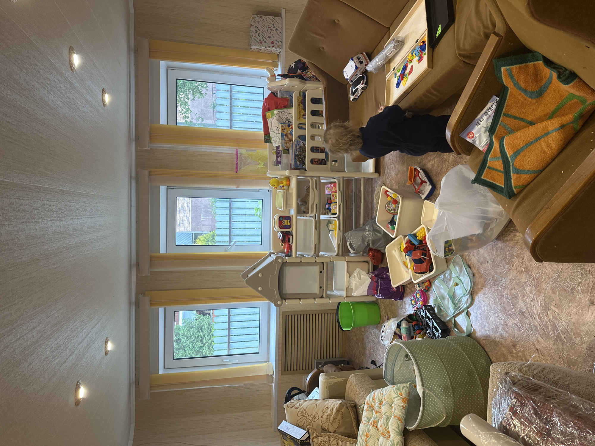 A child is playing with toys in a room with three windows. The room contains two sofas, a white plastic shelving unit, and multiple containers of toys. The location is most likely indoors, possibly in an apartment in Nizhny Novgorod, Russia. The focus is on the child interacting with the toys amidst a cluttered environment. The specific street or building cannot be determined from the image alone.