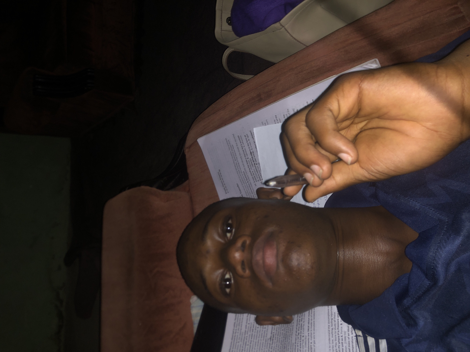 A man with dark skin lies on what appears to be a bed or similar soft surface, next to a table. He's holding a pen over papers covered in printed text. The location is likely indoors. There is no other information to place the image specifically in Atani, Nigeria.