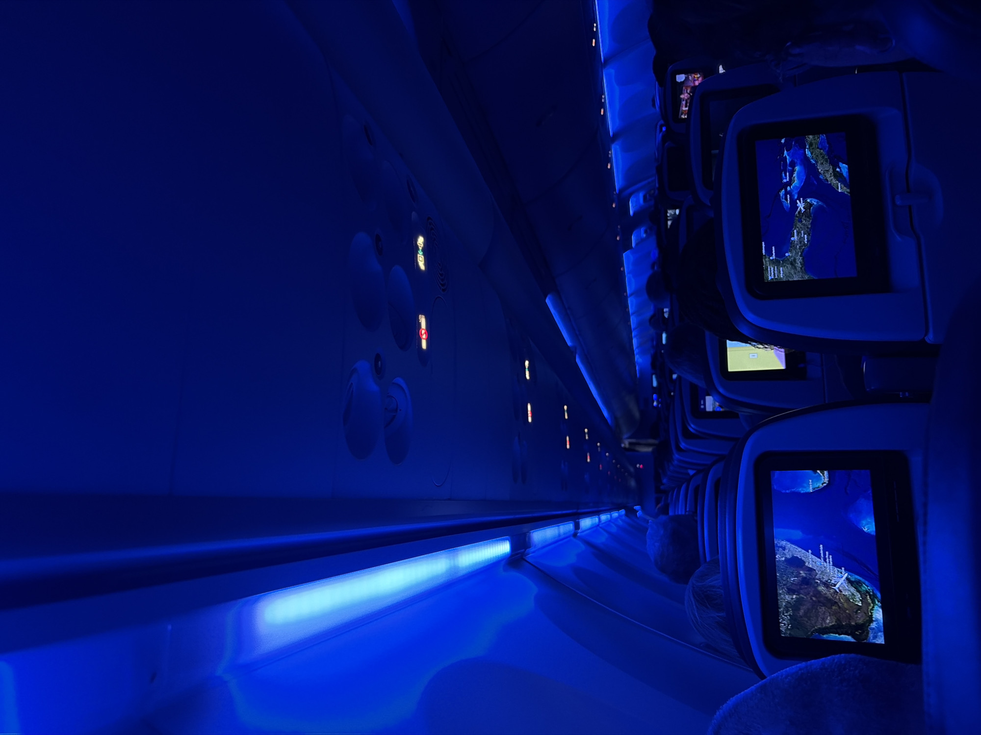 The interior of an airplane cabin is illuminated with a deep blue light. On the right, a row of passenger seats is visible, each featuring a personal entertainment screen. The screens display maps, appearing to show the progress of the flight. On the left, the cabin wall is lined with circular air vents and emergency signage. The overall scene suggests a flight taking place at night or under dimmed lighting conditions. The location cannot be determined solely from the image.