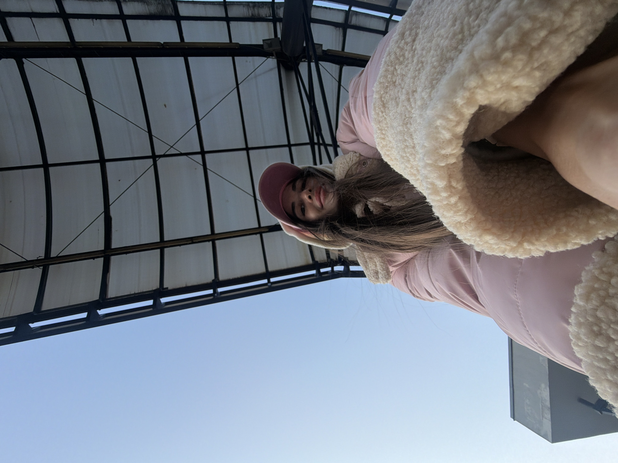 A low-angle shot shows a woman with brown hair, wearing a pink jacket with a beige sherpa lining, and a matching pink cap. The shot is taken from below, looking up at her. Behind her, there's a structured metal framework with a translucent cover, suggesting some kind of roof. The background also includes a clear, blue sky. The building structure is reminiscent of the historical architectural style found in Saint Petersburg, Russia. There is a corner of a modern-looking building on the right side of the shot.