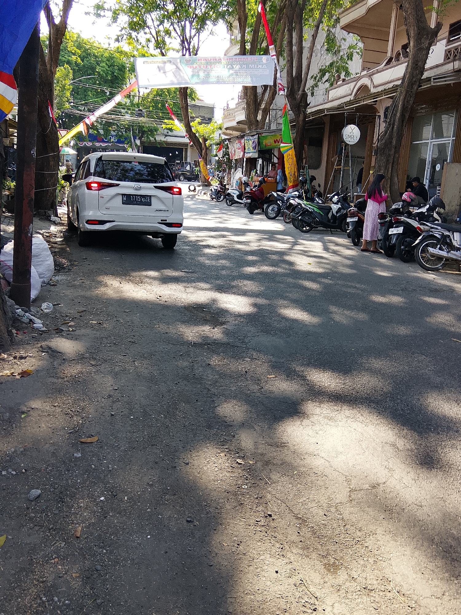 The image depicts a street scene in Kemuteran, Indonesia, with a focus on parked motorcycles and a white SUV. Sunlight casts dappled shadows across the asphalt road, suggesting it is daytime. The white SUV, identified by its license plate "W 1715 ZZE", is parked on the left side of the road, facing away from the viewer.

A row of various motorcycles is parked on the right side of the road. Several individuals are present, including a woman in a pink dress standing near the motorcycles. Another person, wearing a dark shirt, is seated on a motorcycle. Further down the street, more motorcycles and people are visible, along with buildings lining the right side of the road.

The environment includes trees with lush green foliage overhead, some of which are casting shadows. Banners and flags are also visible, strung across the street and attached to poles, indicating a festive or public event. Debris and trash are scattered on the left side of the road, near the parked SUV. The overall impression is of a busy urban street with a strong presence of two-wheeled vehicles.