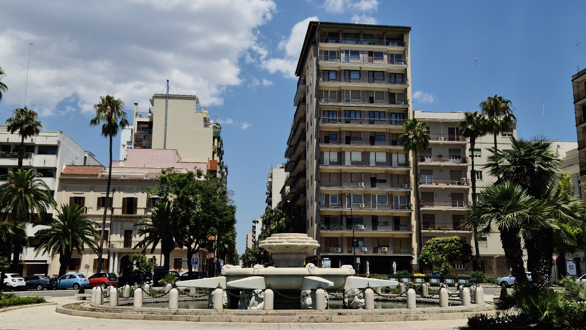 The image shows a sunlit street scene in Taranto, Italy, focusing on a decorative fountain in the foreground and tall buildings and palm trees in the background. The fountain, made of stone, features intricate carvings and is surrounded by a circular paved area. Beyond the fountain, a street is lined with various buildings, including a prominent, multi-story apartment building with numerous balconies and windows. Palm trees of varying heights are interspersed between the buildings, their fronds reaching towards the bright blue sky dotted with fluffy white clouds. Cars are visible parked along the street, and some pedestrians are implied by the presence of sidewalks and shops. The scene conveys a sense of urban life in a warm, Mediterranean climate.