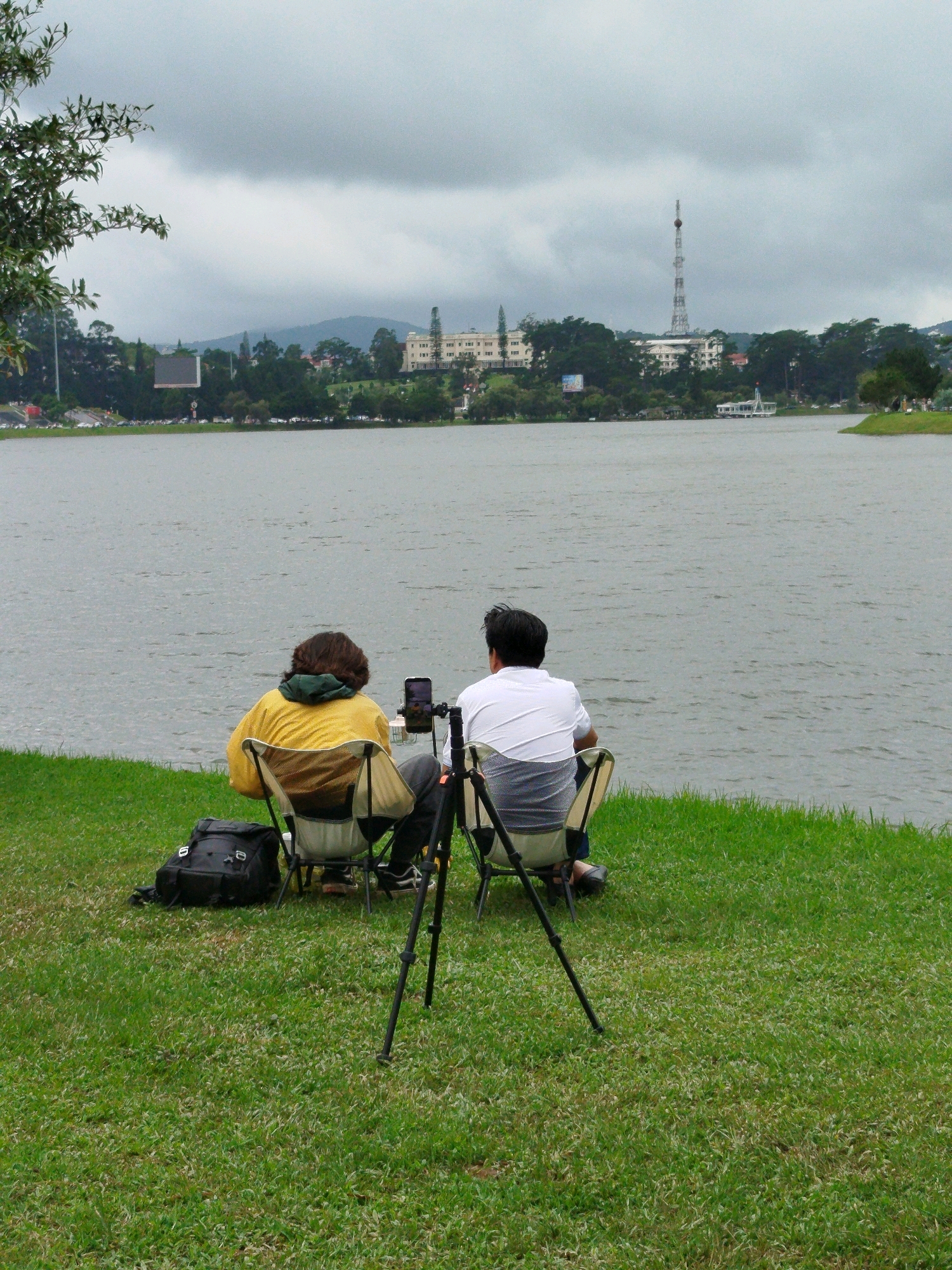 Two individuals are seated on camping chairs facing away from the camera, looking out over Xuan Huong Lake in Da Lat, Vietnam. A black backpack is placed on the grass to the left of the seated figures. A tripod is positioned in front of the individual on the right, with a smartphone attached to its top. The sky is overcast, and the water of the lake appears calm. In the background, buildings and a tall transmission tower are visible on the distant shore, with lush green hills behind them. The scene is captured from a low angle, focusing on the foreground where the people and lake are the primary subjects.