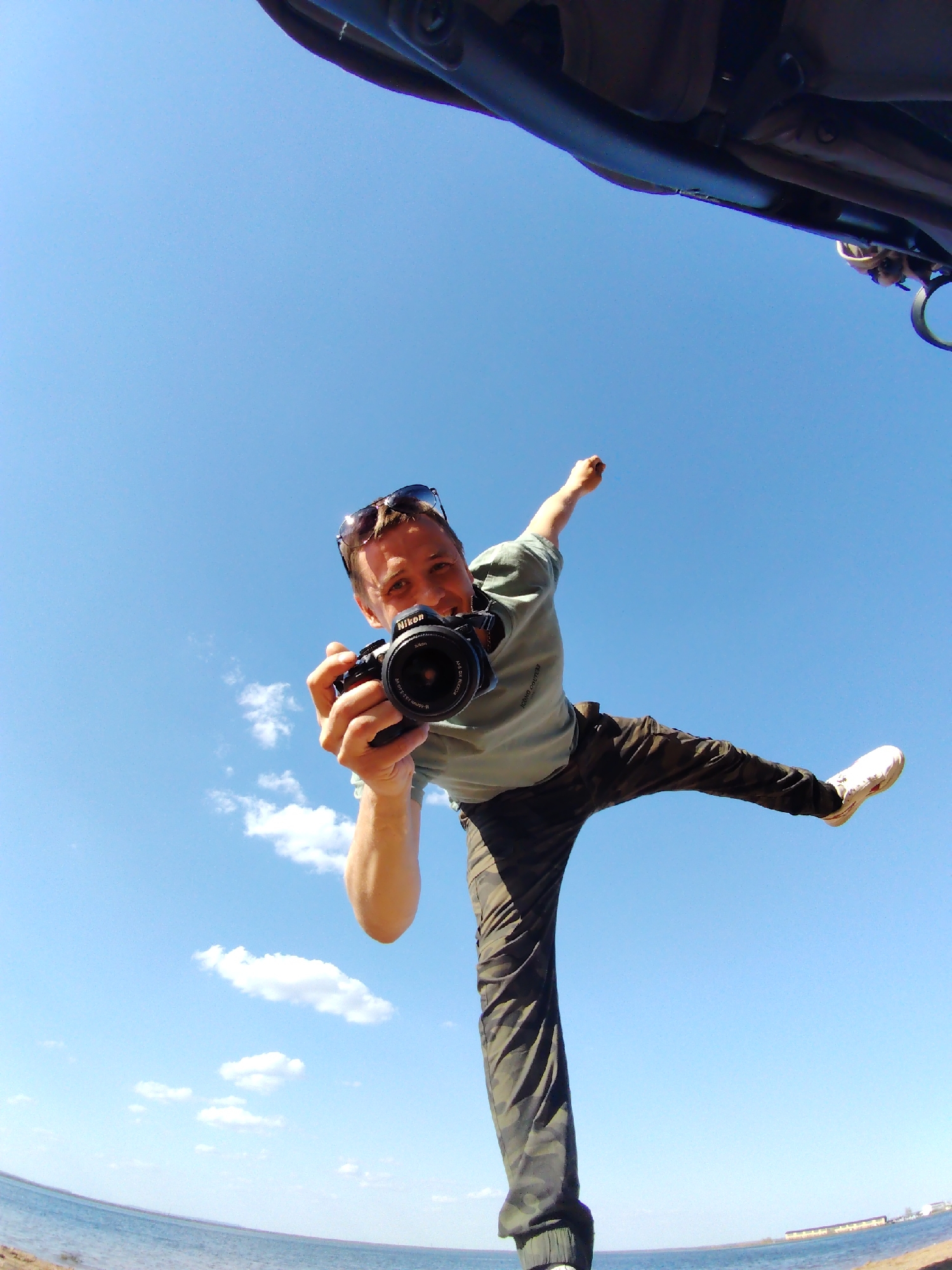 A person is holding a Nikon camera and appears to be in mid-air, against a bright blue sky. They are wearing a light green t-shirt, camo pants, and white sneakers. Sunglasses are perched on their head. In the bottom left corner, there's a glimpse of a shoreline with water and possibly some land or buildings in the distance. The photo was taken in the city of Chaikovsky, Russia. A partial view of what appears to be the inside of a vehicle or structure is at the top of the frame. The overall impression is a dynamic, possibly humorous, self-portrait in an outdoor setting.