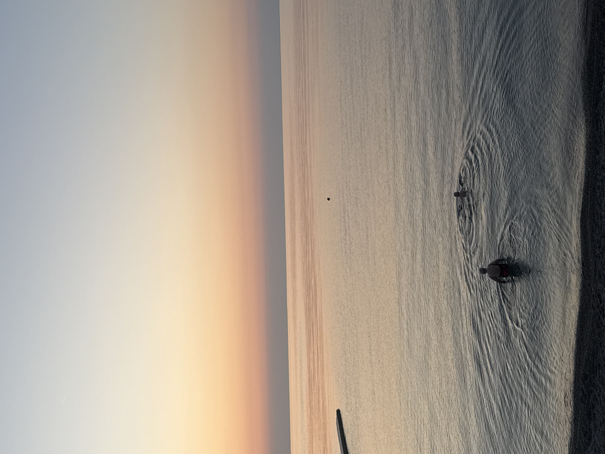 The image shows a serene seascape at dusk, with two individuals partially submerged in the calm waters of the Black Sea. The sky displays a gradient of colors, transitioning from a pale blue at the top to soft shades of peach and pink near the horizon, suggesting the setting sun. A crescent moon is faintly visible in the upper left portion of the sky. The water is smooth with subtle ripples, reflecting the muted tones of the sky. In the foreground, a person is standing in the water, facing away from the viewer, with their upper body visible. Further out in the water, another individual is swimming, leaving a wake behind them. A narrow, dark structure, possibly a pier or breakwater, extends into the water from the left side of the frame. The shoreline in the very bottom is composed of dark pebbles. The overall scene evokes a sense of tranquility and solitude. The location is specified as Partenit, Ukraine.