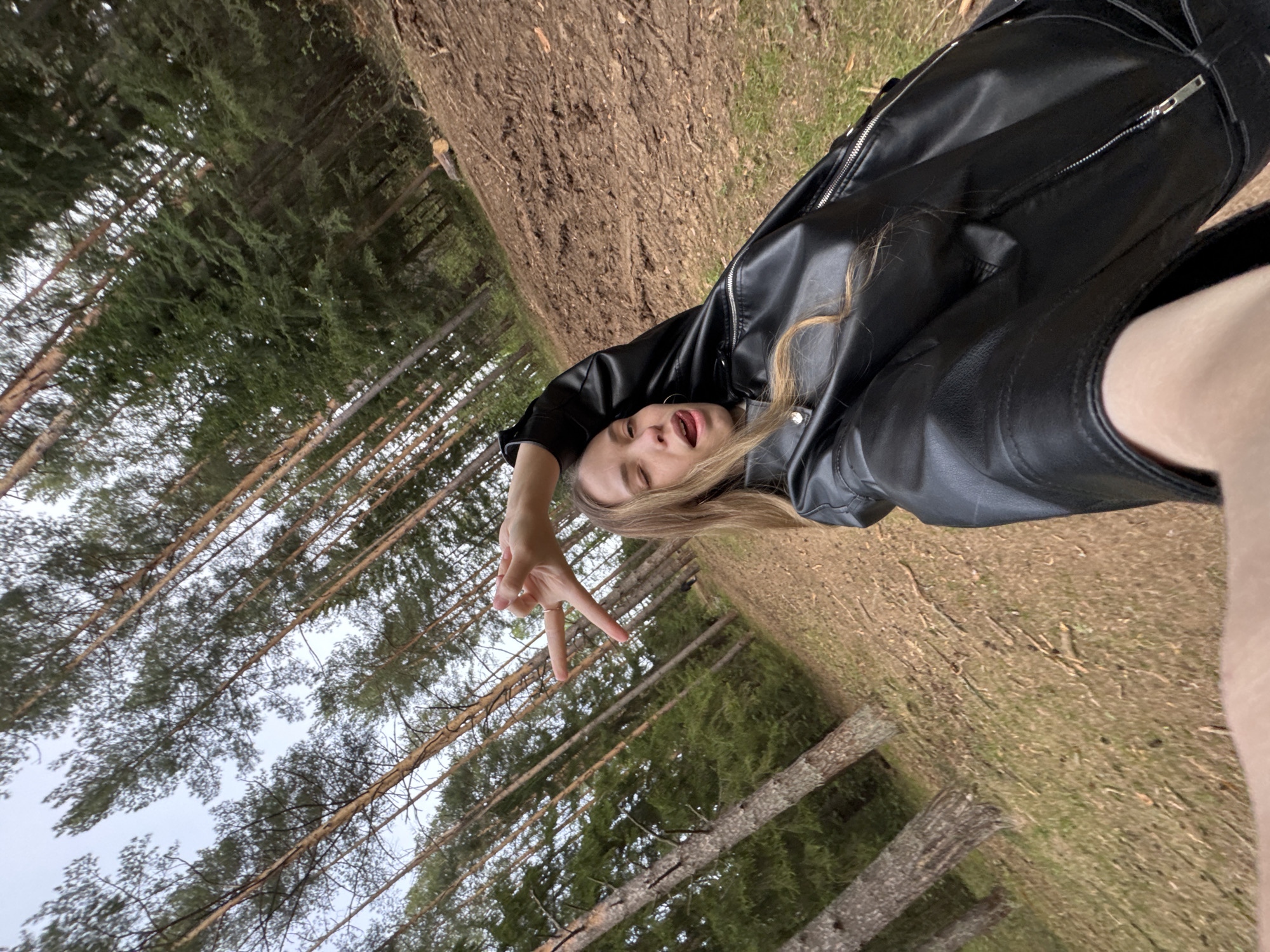 A young woman is lying on her side in a forest, making a peace sign with her right hand. She is wearing a black leather jacket. The background consists of tall trees and grass. The location is outdoors, likely in a forest area near Прикамский, Russia, given the context.