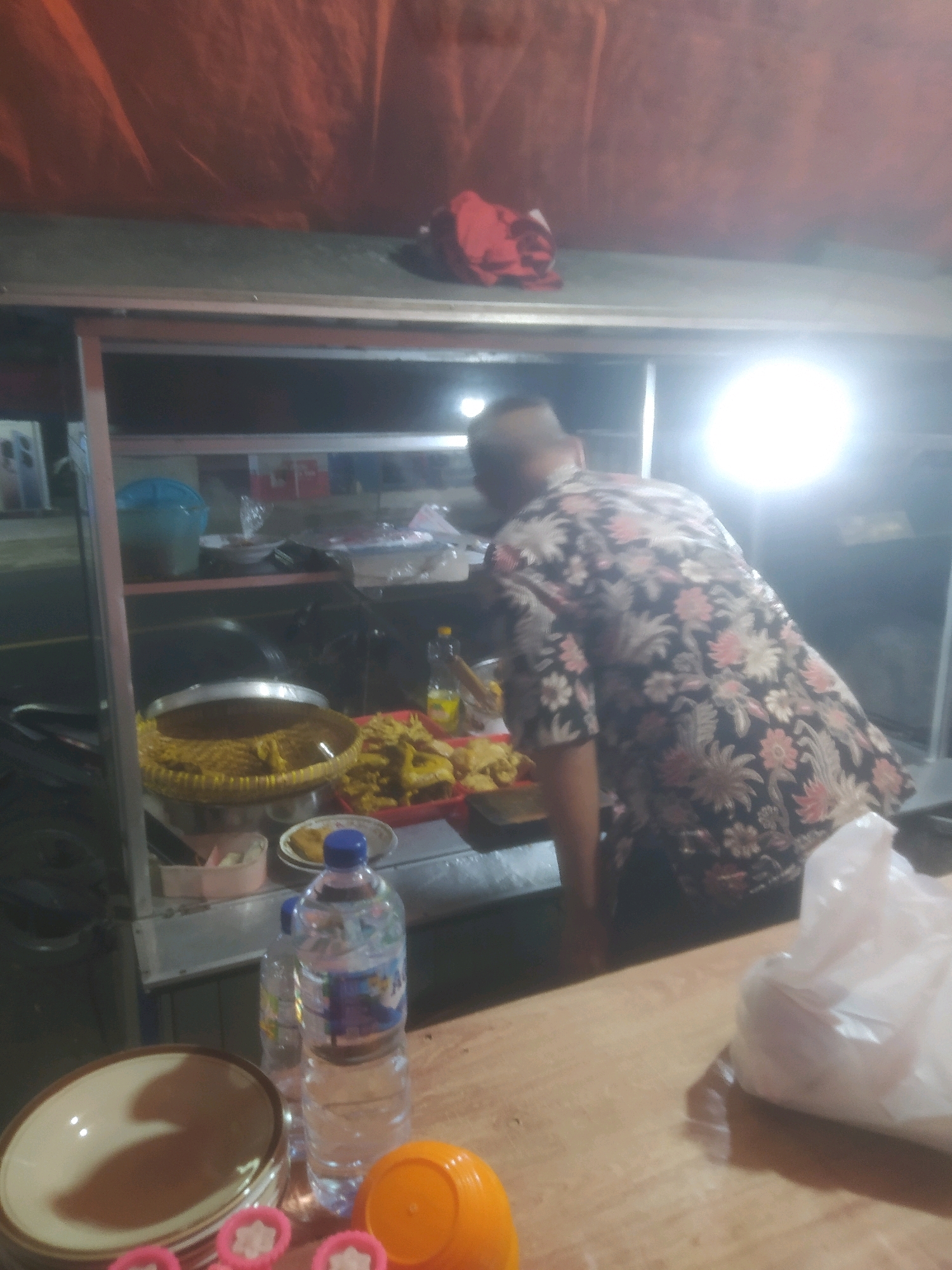 A man in a floral shirt is standing behind a food stall in Desa Karangbenda, Indonesia. He appears to be preparing or serving food. Various food items, including fried foods and noodles, are displayed on the stall. Bottles of water and other containers are also present. The stall is covered by an orange tarp and illuminated by bright lights, suggesting it is nighttime or a dimly lit area. The overall environment appears to be an outdoor market or street food vendor.