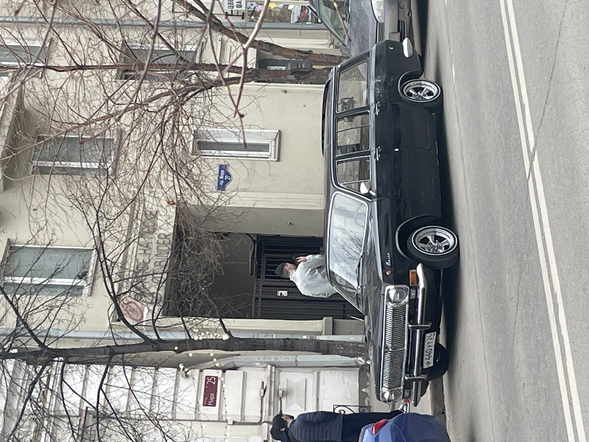 A black GAZ-24 Volga station wagon is parked on the side of a street in Krasnoyarsk, Russia. The car has aftermarket wheels. A man in a light-colored jacket is standing by the entrance to a building. Two people are standing near the front of the Volga. The building behind the car has a sign on it in Russian. A street sign shows the number 4. A sign indicating "Mira 25" is visible on a building. The environment appears to be urban, and the trees are bare, suggesting it is autumn or winter. The license plate on the Volga appears to be P440YT24.