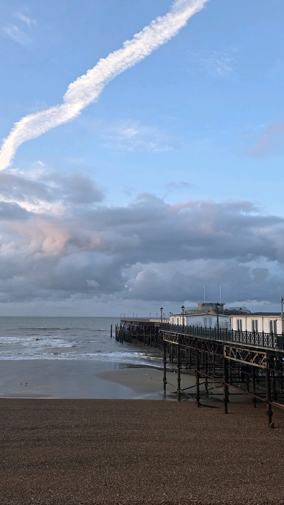 The image depicts a view of the St Leonards Pier in St Leonards-on-Sea, United Kingdom. The sky is partially cloudy with a distinct contrail of an aircraft visible in the upper left quadrant, curving across the blue sky. Below the sky, a line of grey clouds stretches across the horizon, with hints of pinkish light reflecting from the setting or rising sun.
The sea is visible, with moderate waves breaking on a sandy and pebbly beach. The tide appears to be low, revealing wet sand. Several small birds are scattered on the wet sand.
In the foreground and right side of the image is the St Leonards Pier. It is a substantial wooden and metal structure extending out over the water. The pier has a railing with ornate metalwork and is lined with lampposts. On the pier, there are several white buildings with windows, likely housing shops or attractions. The underside of the pier shows a complex structure of wooden beams and metal supports. The beach in the foreground is covered in small, dark pebbles.
There are no people visible in the image. The overall atmosphere suggests a cool, possibly overcast day at the seaside.