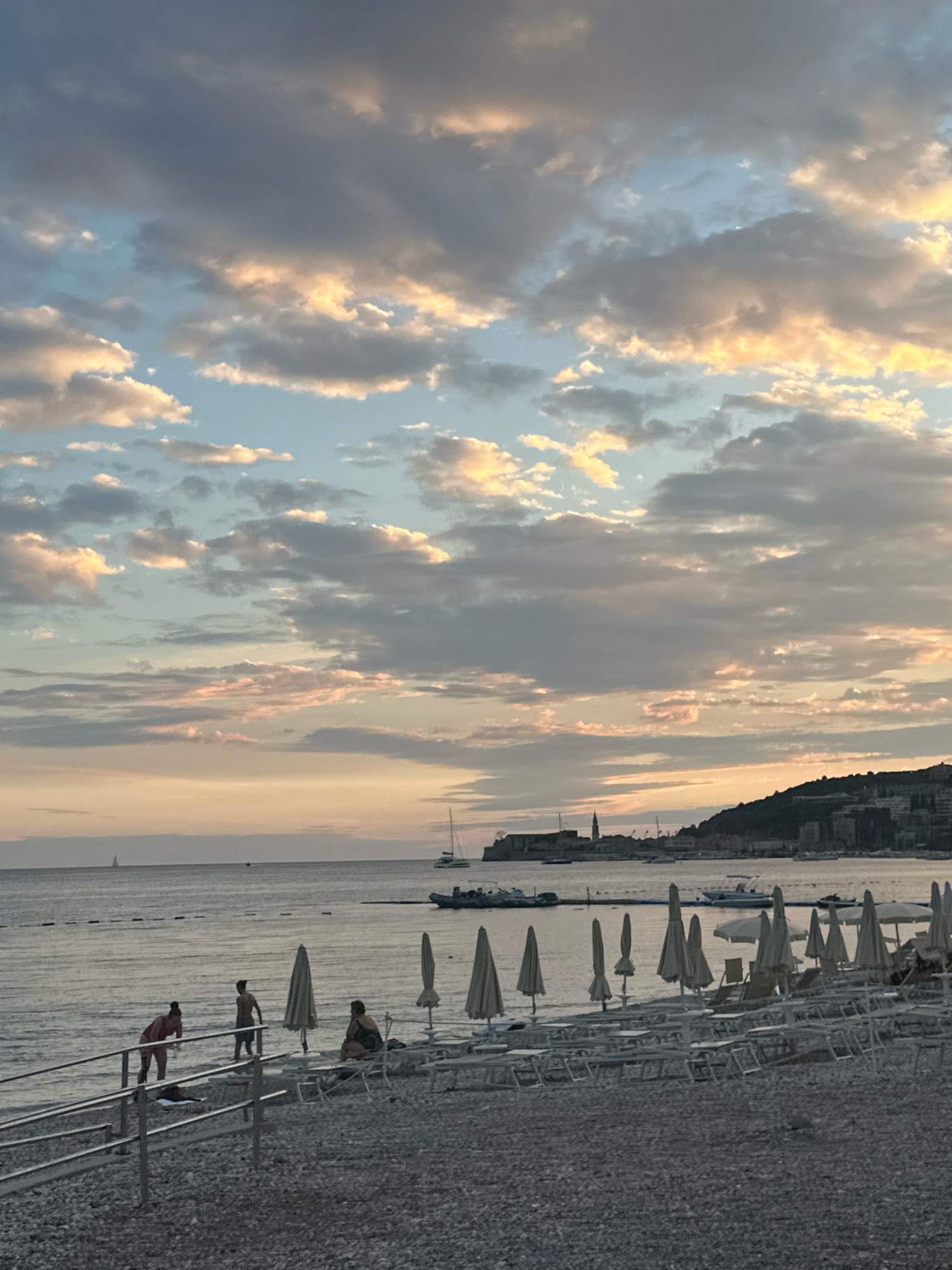 The image depicts a tranquil beach scene in Budva, Montenegro, during what appears to be sunset or sunrise. The sky is filled with dramatic clouds, tinged with soft hues of orange, pink, and blue. In the distance, a coastal city skyline is visible, with buildings and a prominent church tower nestled on a hillside. The sea is calm, with a few sailboats and a motorboat on the horizon.
On the pebble beach in the foreground, rows of closed beach umbrellas and empty lounge chairs are neatly arranged, suggesting the end of a day at the beach. A few individuals are present near the water's edge. Two people, one wearing a red top and shorts, the other in dark shorts, are standing by a metal railing leading down to the water. Another person, wearing dark shorts, is walking away from the railing and towards the beach. A fourth person is seated on a lounge chair further back on the beach. The overall atmosphere is peaceful and serene.