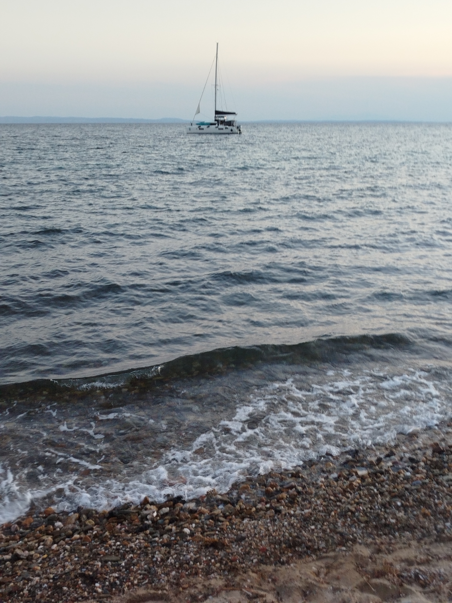 A wide shot captures a sailboat on a calm sea, with a pebbly beach in the foreground. The sky is a hazy gradient of pale blue and gray, suggesting either early morning or late evening. The sailboat, white with a black hull and a single mast, is positioned in the middle ground, appearing to be at anchor or moving slowly. The sea is a deep, undulating blue, with gentle waves lapping at the shore. The beach is composed of a variety of small, smooth pebbles in shades of brown, gray, and white, with a strip of sand visible at the very bottom of the frame. The overall scene is serene and tranquil, evoking a sense of peacefulness. The location is likely a coastal area within the Sithonia Municipal Unit, Greece, given the characteristic landscape. There are no people visible in the image.