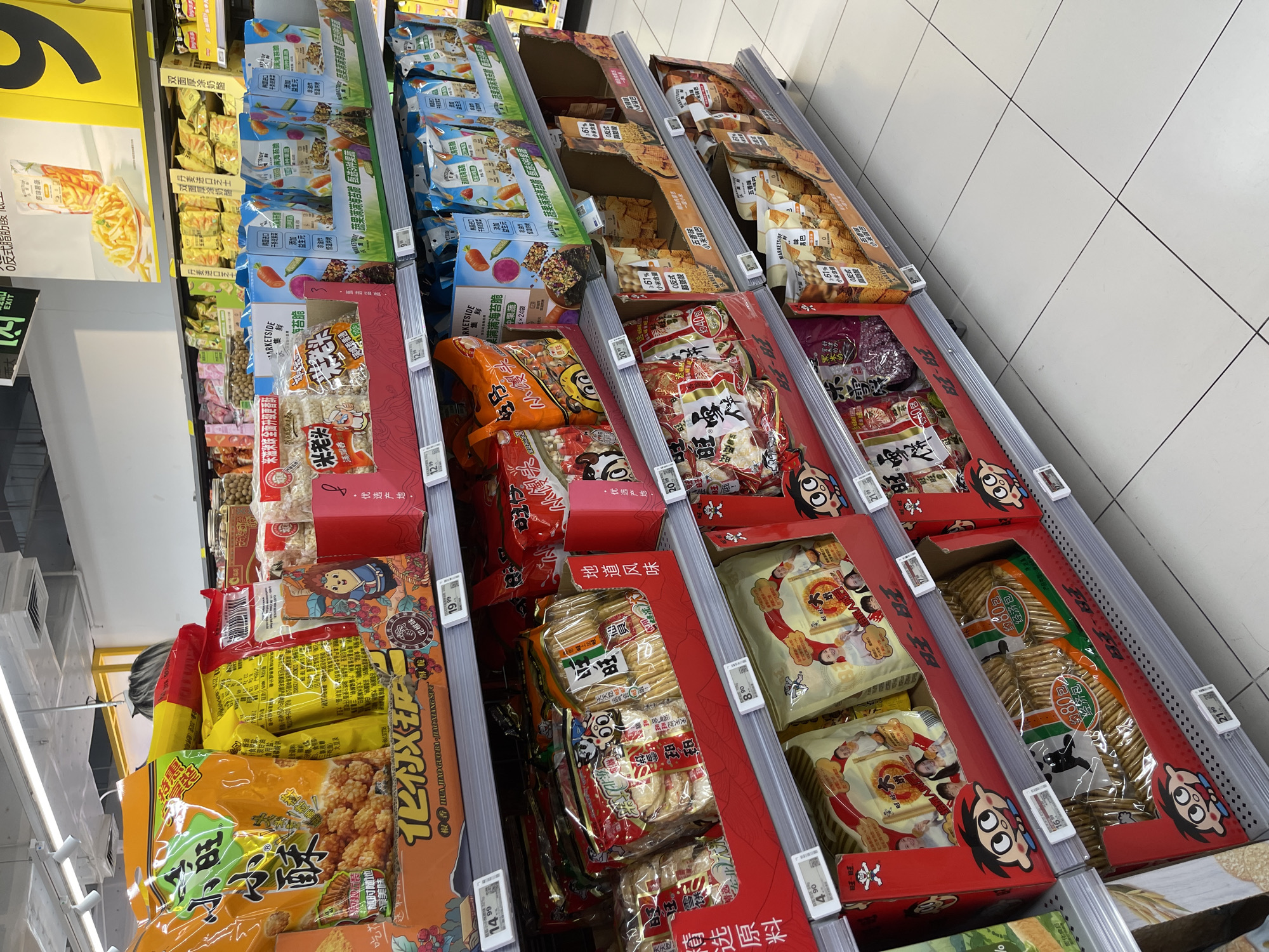 This is a medium shot of a supermarket aisle in Panyu District, China, displaying shelves stocked with various packaged snacks. The focus is on the snacks themselves, which are predominantly in colorful bags and boxes. The shelves are organized and well-lit, with price tags visible in front of some products. The merchandise includes items like puffed rice snacks, crackers, and other dry snack foods, many with Chinese text on their packaging. The overall environment is typical of a modern grocery store, with clean white tiled flooring and overhead lighting fixtures. There are no people actively browsing or interacting with the products in the frame. The scene is static and focuses on the commercial display of goods.