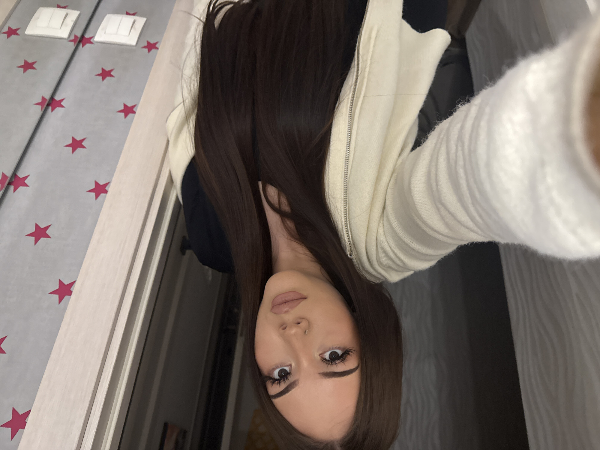 The image, rotated 90 degrees counter-clockwise, presents a close-up indoor selfie of a young woman, likely taken within a residence in Naberezhnye Chelny, Russia.
**Subjects:** The primary subject is a young woman with long, straight, dark brown hair. She looks directly into the camera with a neutral expression. Her makeup includes defined eyebrows, long, dark eyelashes, and nude-toned lipstick. A small stud piercing is visible on her nose.
**Clothing:** She is dressed in a cream or off-white zippered hoodie or jacket, worn over a dark top. The fabric of the hoodie appears soft, possibly knitted.
**Scene and Setting:** The background on the left side of the image features a light gray or off-white wall adorned with a repeating pattern of bright red stars. This star-patterned surface appears to be a mirror, reflecting parts of the room. Two white rectangular light switches are mounted on this mirrored wall. Adjacent to the mirrored surface, a light-colored wooden door frame or trim is visible, suggesting an architectural opening. The right side of the image shows a darker, less distinct area of the room, possibly another wall with subtle vertical texturing. The lighting suggests an interior setting, likely illuminated by artificial light.
**Activity:** The woman is posing for a self-portrait, holding the camera at a slightly elevated angle.
**Notable Details:** The distinct red star pattern on the mirrored wall provides a personal decorative element to the room. There is no visible text in the image. The time of day is not clearly indicated by natural light, suggesting an indoor setting with artificial illumination.