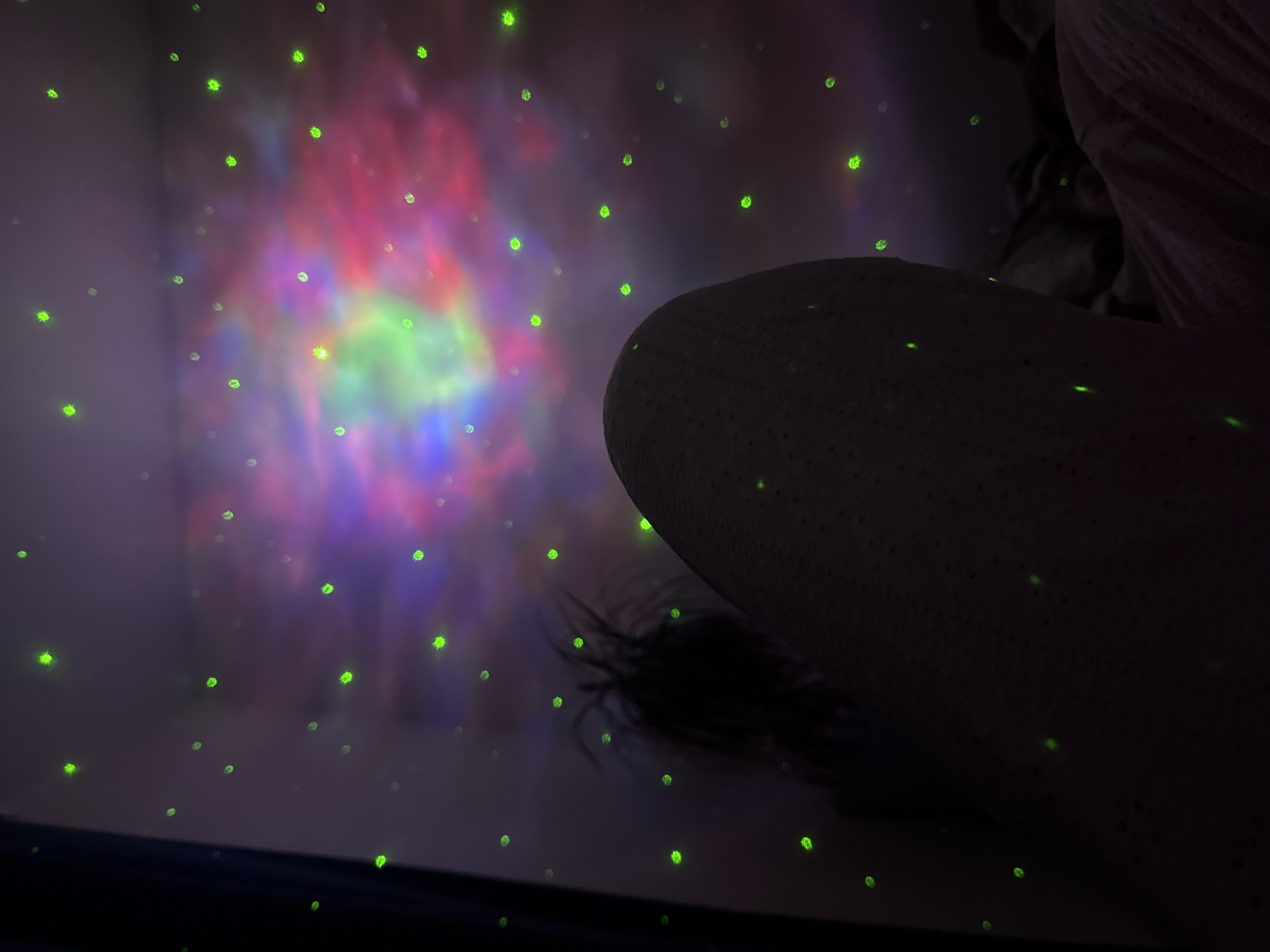 The image depicts a dimly lit room with a projection of colorful nebulae and green lights resembling stars on the wall and ceiling. In the foreground, the side of a person's body, possibly a leg, is visible, covered in a textured material. To the left, a dark silhouette of a plant with spiky leaves is partially visible. The overall impression is of a relaxed, possibly nighttime setting, perhaps in a bedroom, with ambient lighting creating a celestial atmosphere. The image does not provide any information that can be used to determine its location in the city of Perm, Russia.