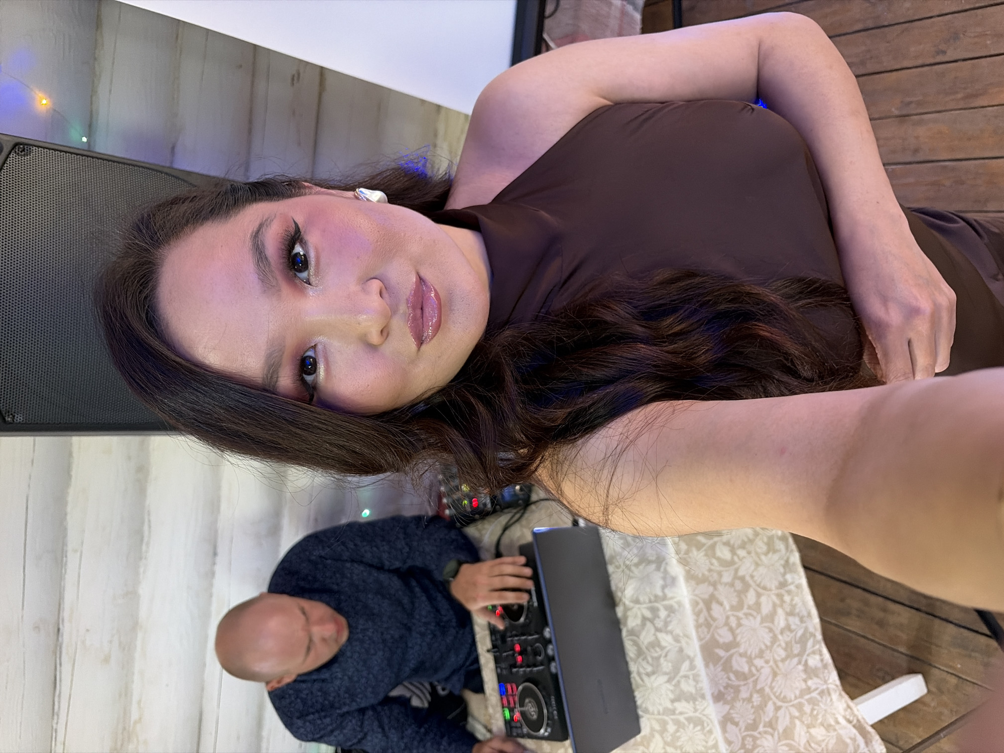 This media file captures a selfie of a woman taken indoors, featuring a man operating DJ equipment in the background. The woman, prominently in the foreground, has long, wavy dark hair and is wearing a dark brown, possibly one-shoulder or halter-neck, top. She looks directly at the camera with a neutral expression, sporting winged eyeliner, pink eyeshadow, and glossy lips. A white earbud is visible in her right ear.
In the background, a bald man in a dark blue patterned sweater is seated, focusing on a DJ mixer and a laptop placed on a table. The table is covered with a cream-colored tablecloth featuring a subtle floral pattern. This setup suggests an event or party where the man is acting as a DJ.
The setting includes light-colored wooden paneling or planks, possibly a wall or floor, and a large black speaker on the left side of the frame. Small, colorful decorative lights can be seen in the upper left background, indicating an evening event or a venue with mood lighting. The overall lighting is dim, contributing to a nighttime atmosphere. The image is taken from a slightly tilted perspective, with the woman's head angled towards the camera. No discernible text is clearly visible on any objects. The scene is located in what is contextually provided as Прикамский, Russia.