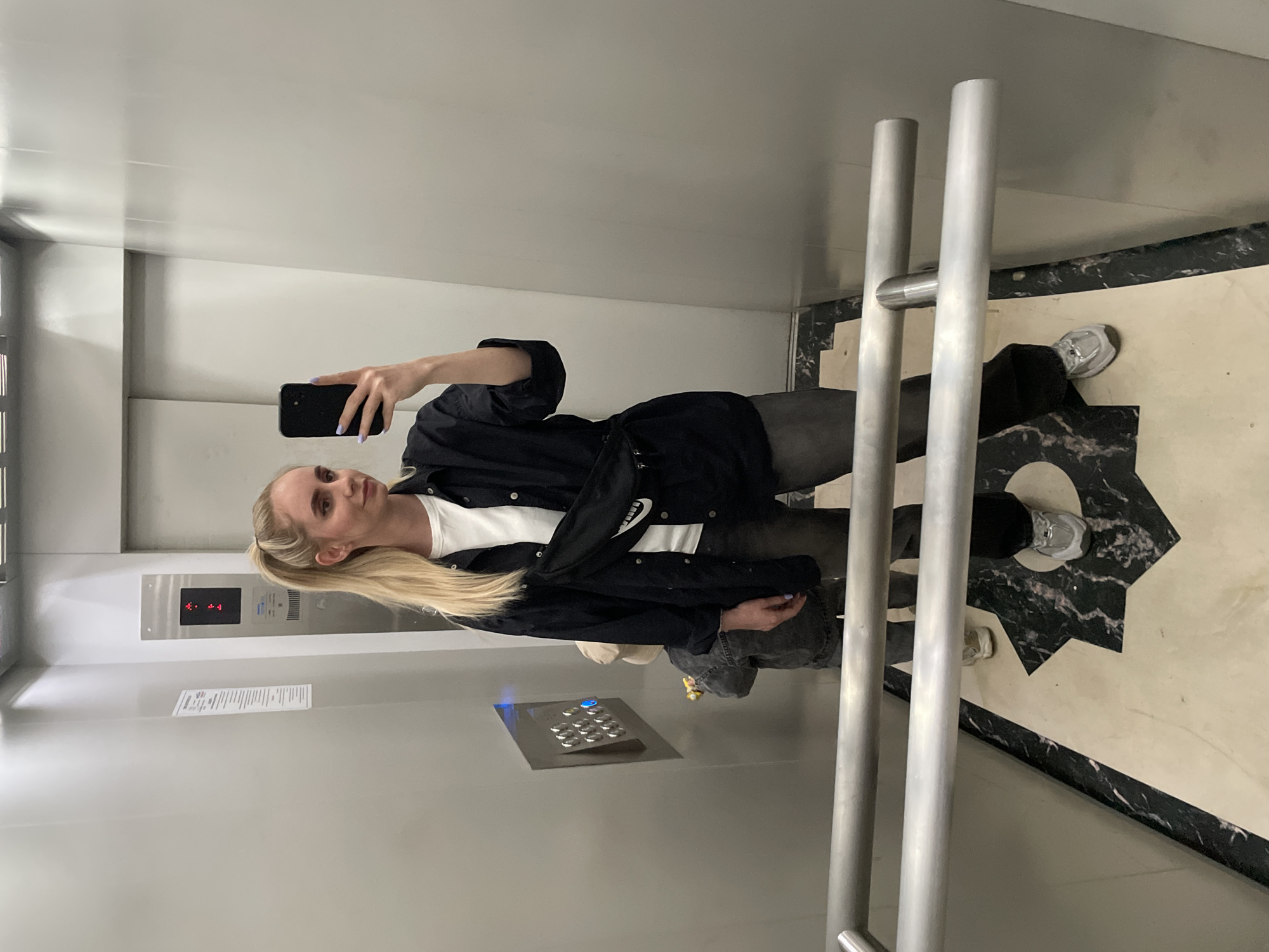 A woman is taking a selfie in an elevator. She is fair-skinned with long blonde hair pulled back in a ponytail. She is wearing a white t-shirt, a black jacket, grey pants, and silver sneakers. She is holding a black smartphone in her left hand with lavender nail polish. The elevator has stainless steel walls and a panel of elevator buttons. The floor is decorated with a black and white geometric pattern. A handrail is present in the foreground. The image was taken in Vladivostok, Russia.