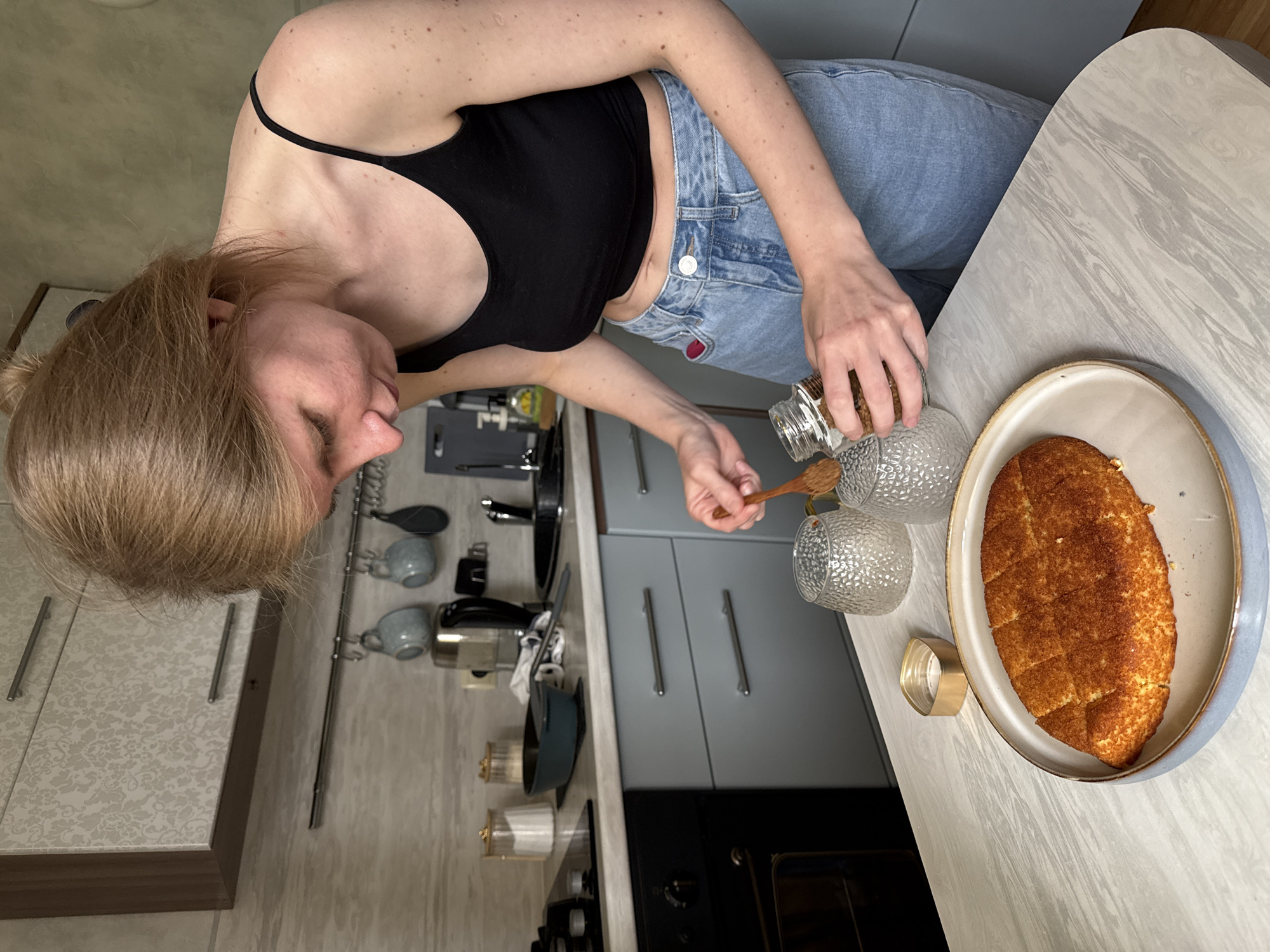 A woman with blonde hair, wearing a black tank top and blue jeans, stands at a table in a kitchen, possibly in Izhevsk, Russia. She is using a wooden spoon to take something from a glass jar and place it into one of two textured glass cups. Next to the cups is a plate containing a golden-brown, sliced loaf of bread. The kitchen is fitted with gray cabinets and drawers with silver handles. Cooking utensils hang on the wall. An oven and stove are visible below the countertop. The table is a light gray color, with a white marbling pattern.