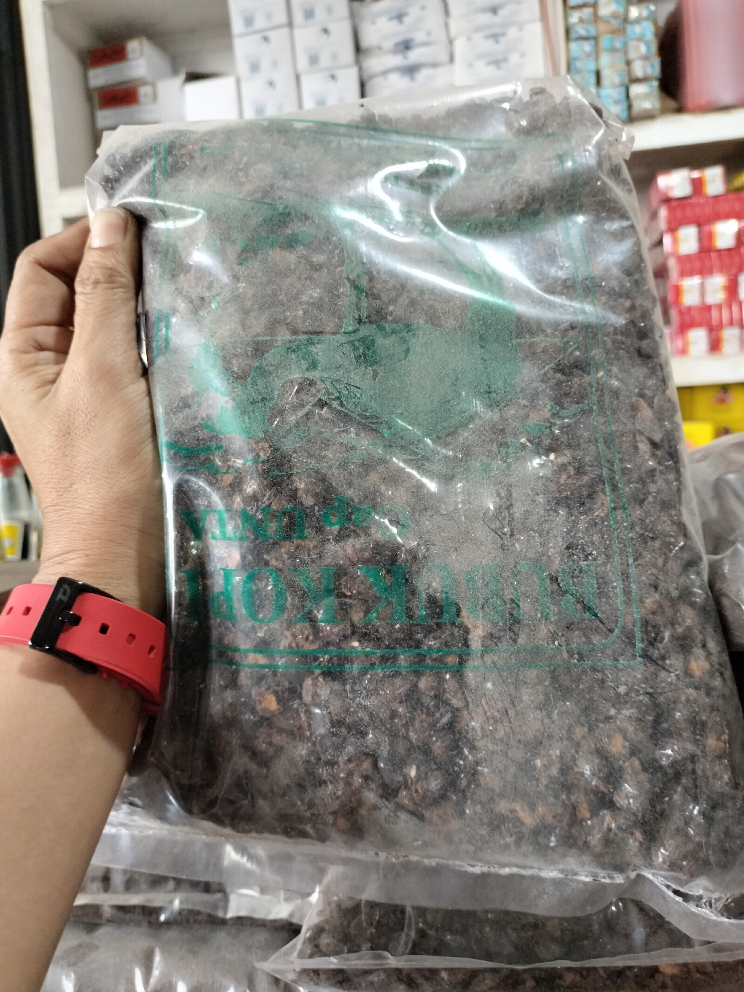 A person's left arm and hand are holding a clear plastic bag filled with dark brown, granular material. The bag has green lettering that appears to be a product label. The person is wearing a red wristwatch on their left wrist. The background shows shelves stocked with various items, suggesting an indoor retail environment, possibly a shop in Kotapantonlabu, Indonesia.