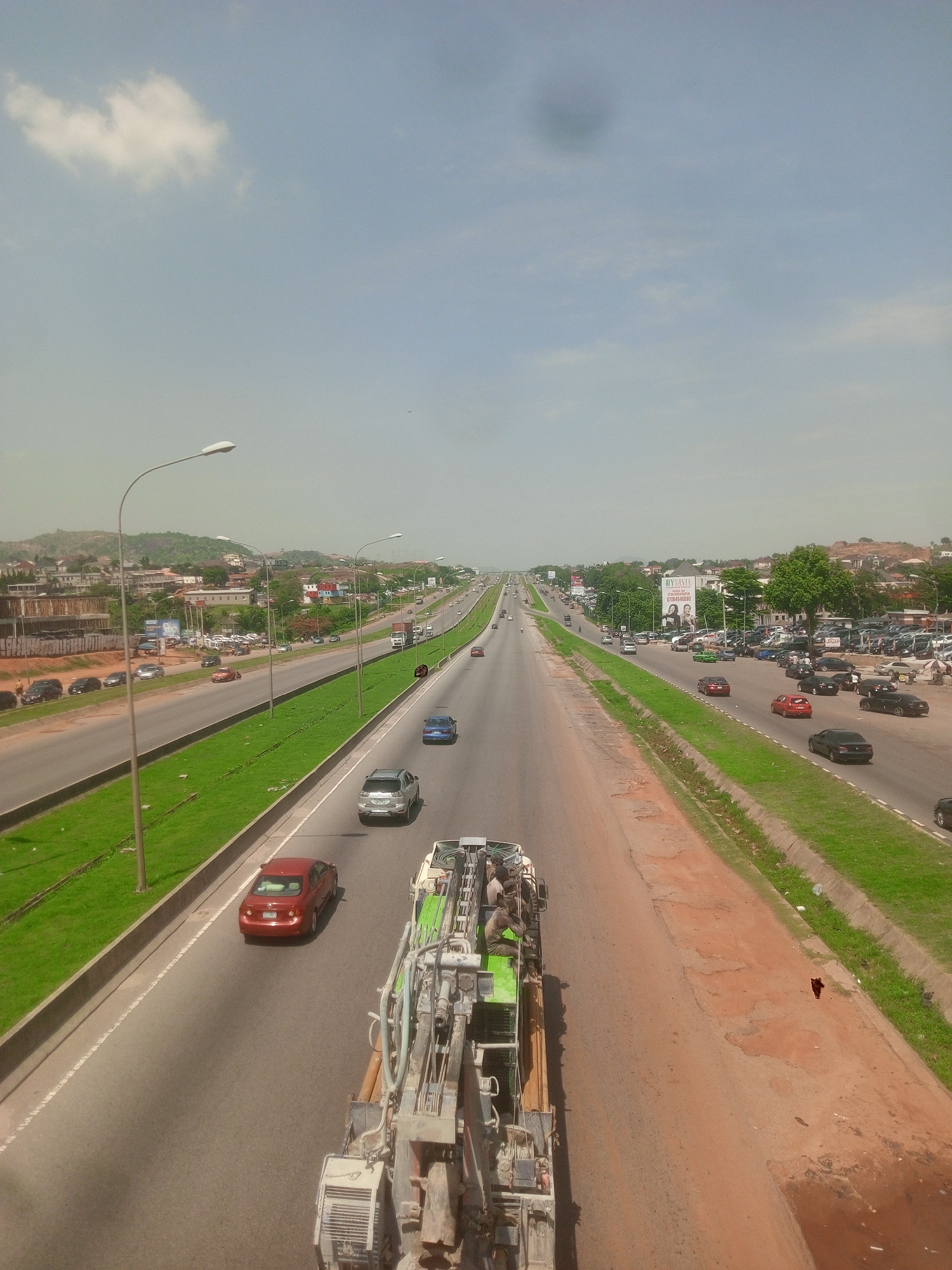 The image depicts a multi-lane highway in Unknown, Unknown. The roadway is filled with various cars, including a red sedan and a silver sedan. A specialized work vehicle is in the foreground, carrying several individuals. The highway is bordered by green medians with street lamps. In the background, buildings and trees are visible, indicating a city environment. The sky is pale blue with light cloud cover.