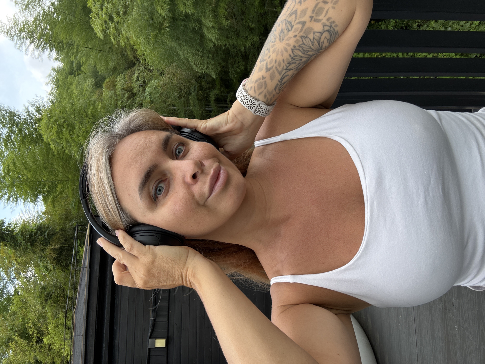 A woman with blonde hair and blue eyes is pictured wearing a white tank top and black headphones in a natural setting. She is holding the headphones to her ears with both hands. Her left arm is adorned with a tattoo of floral designs and a white bracelet. She is standing outdoors with lush green trees and bamboo in the background. A dark-colored structure with a railing is visible to her left. The location is likely a residential area or a recreational space, possibly within Wuxing District, China, given the vegetation.