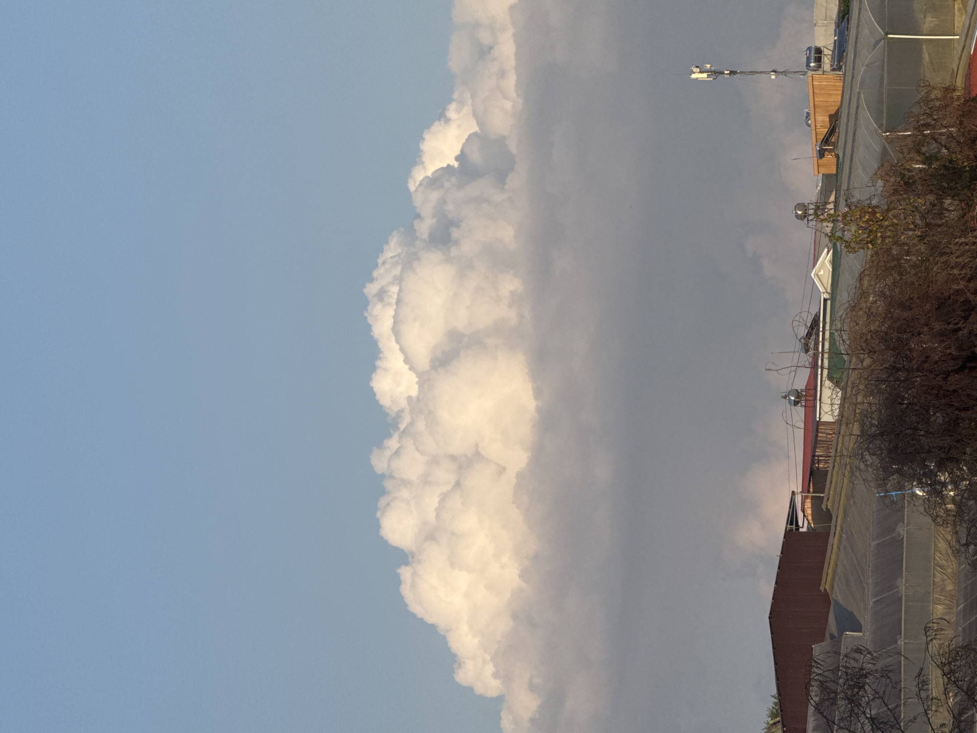 This is a vertical, eye-level, full shot of a cloudy sky over a town in Lâm Viên - Đà Lạt, Vietnam. The sky takes up the top two-thirds of the image, with a large, bright cloud formation dominating the center. The cloud is lit from above, with the top appearing fluffy and white, while the bottom is a darker gray, suggesting it is lower in the atmosphere or blocking light from the sun. Below the clouds, the town's rooftops are visible. These include several structures with corrugated metal roofs, some appearing to be greenhouses with translucent coverings, and other buildings with more traditional roofing. A tall cell tower with antennas stands prominently on the right side of the frame, among the buildings. There are no people or animals visible in the image. The overall scene conveys a daytime atmosphere with dramatic cloud formations.