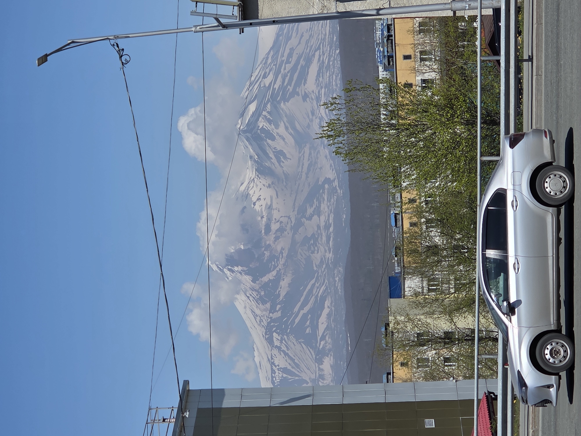 An eye-level, medium shot captures a silver sedan driving across a roadway in front of a backdrop of a snow-capped mountain range, residential buildings, and a bright blue sky. The car, seen from its left side, is positioned in the foreground, slightly to the left of the frame. The mountain range, covered in patches of snow and rock, dominates the background, with wispy clouds clinging to its peaks. Below the mountains, a cluster of colorful buildings, with yellow and white facades and dark roofs, is visible. The scene is further framed by a metal railing in the midground and power lines crisscrossing the sky. A lamppost stands on the right side of the image, with a bicycle chained to the wires. The environment appears to be a city with a prominent natural landmark in the distance.