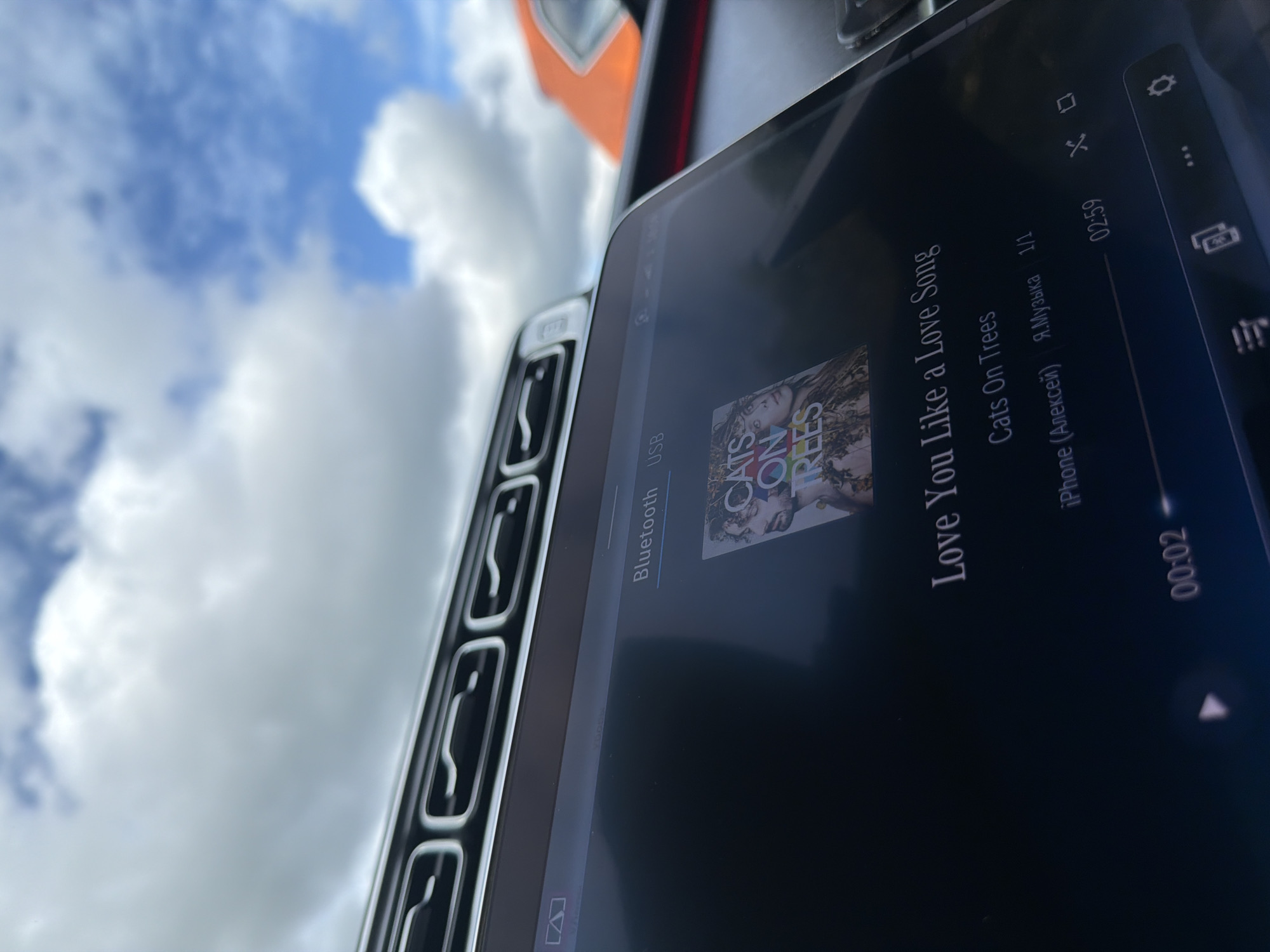 A close-up shot depicts a screen displaying the song "Love You Like a Love Song" by Cats On Trees, sourced from an iPhone via the Я.Музыка platform, playing within a car interior. The song is 2 seconds into a total length of 2 minutes and 59 seconds. Visible car elements include the air vent and portions of the dashboard. The background features a sky filled with clouds. There is no information about the location in the city of Borki, Russia.