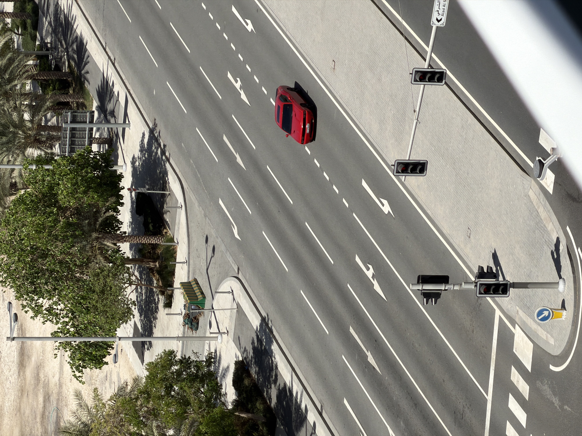 An aerial, high-angle shot captures a wide, multi-lane road with a red car driving on it. The road is paved with asphalt and marked with white lane lines and directional arrows. On the left side of the road, there are trees, a sidewalk, and a street light. On the right side of the road, there is a sidewalk and more street lights, some of which are traffic signals. A sign with Arabic text and "Alad Al S" is visible on the right. The environment appears to be urban, with buildings or structures visible in the background, partially obscured by trees. The sky is not visible in the frame.