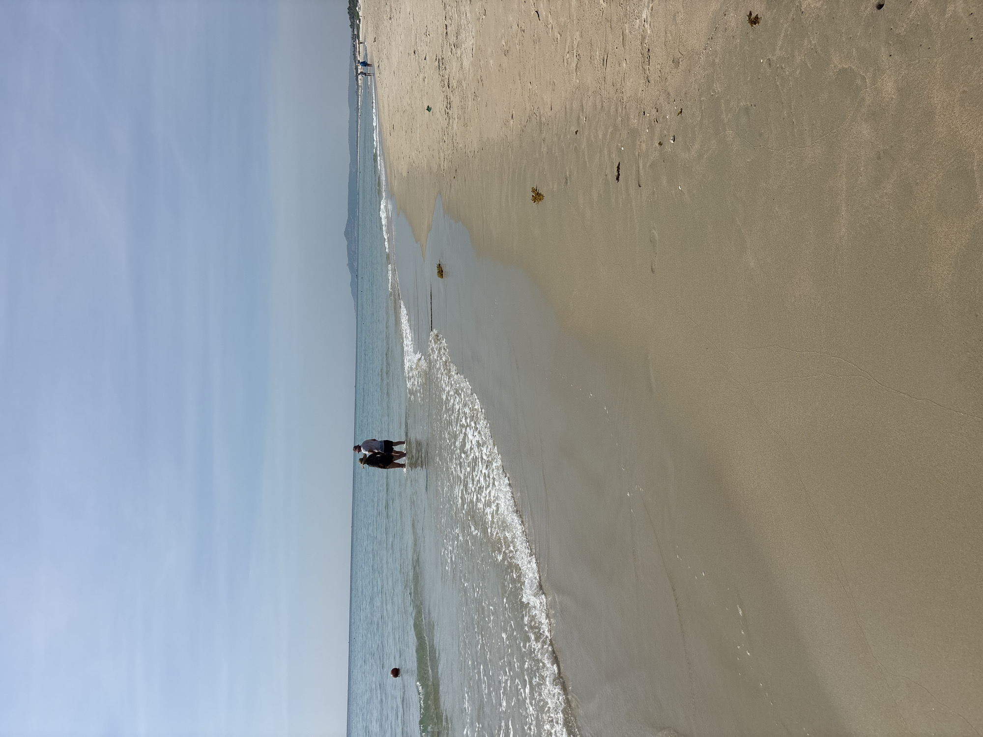 Two individuals are standing in the shallow water of a beach. The person on the left is wearing a dark-colored outfit and a hat. The person on the right is wearing a light-colored shirt and dark shorts. A red spherical object is visible in the water to the left of the two individuals. The beach has light-colored sand with some scattered seaweed. The ocean is calm with gentle waves reaching the shore. In the background, the coastline extends into the distance with hills or mountains. The sky is clear and blue with some wispy clouds. Several other people are visible in the distance along the beach.