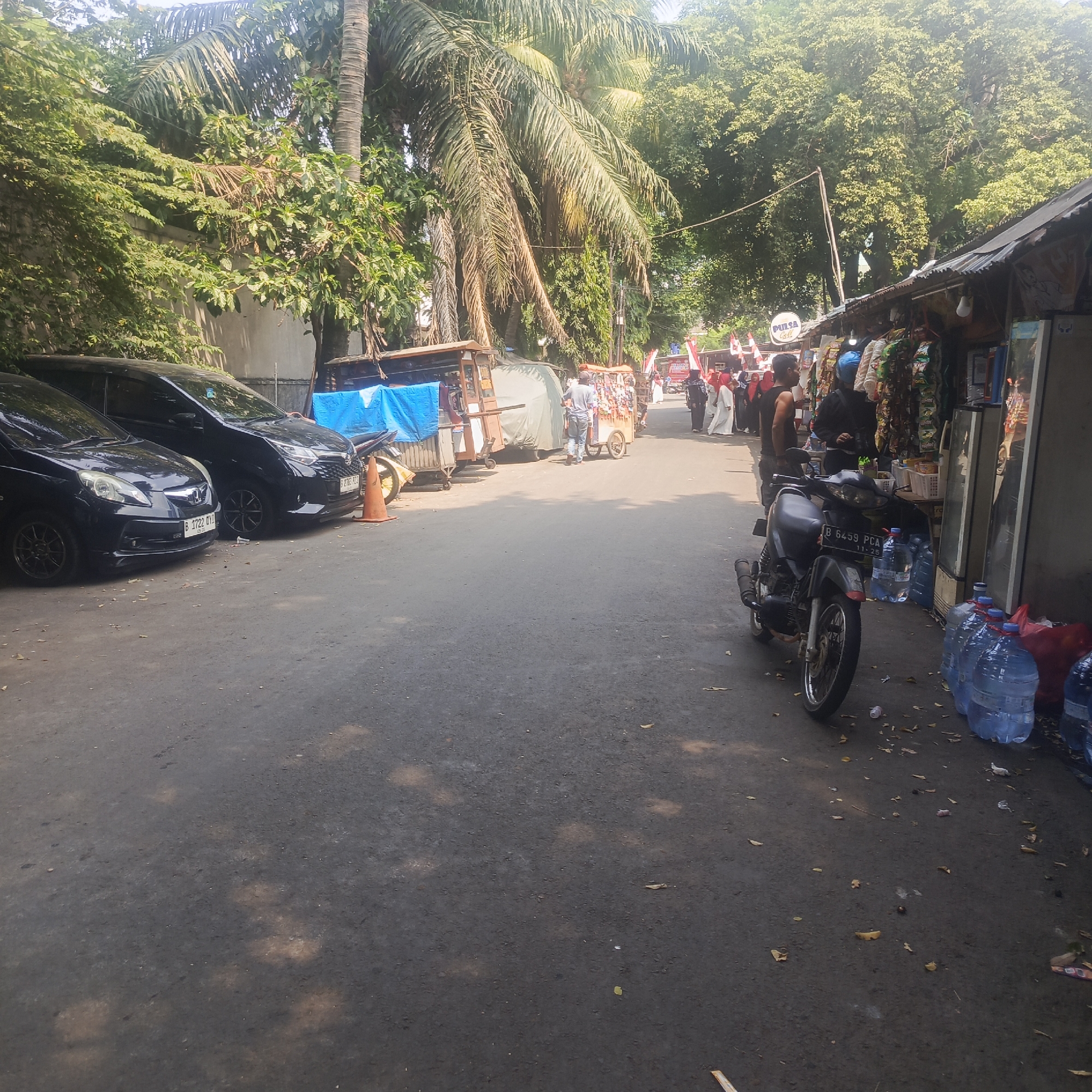 The image depicts a street scene in South Jakarta, Indonesia, likely on a sunny day given the shadows. The foreground is dominated by an asphalt road with fallen leaves and some scattered litter. To the left, two dark-colored cars are parked, one in front of the other. The first car, a hatchback, has a license plate reading "B 1722 OVY". Behind it is a minivan or SUV with license plate "B 2765 PZX".
Further down the street, several vendors are set up along the right side. A motorcycle, with license plate "B 6459 PCA", is parked near a stall. This stall appears to be selling various goods, with rows of items hanging and displayed, and large bottles of water stacked nearby. Other makeshift stalls or carts are visible further down the street.
Several people are present. Some are walking along the street, including individuals dressed in traditional Islamic attire, such as women wearing hijabs. One person appears to be operating a cart or stall. Red and white Indonesian flags are visible, suggesting a national celebration or event. Trees line the background, providing shade and contributing to a somewhat tropical environment. The overall impression is of a busy, everyday street scene with commercial activity and local residents.