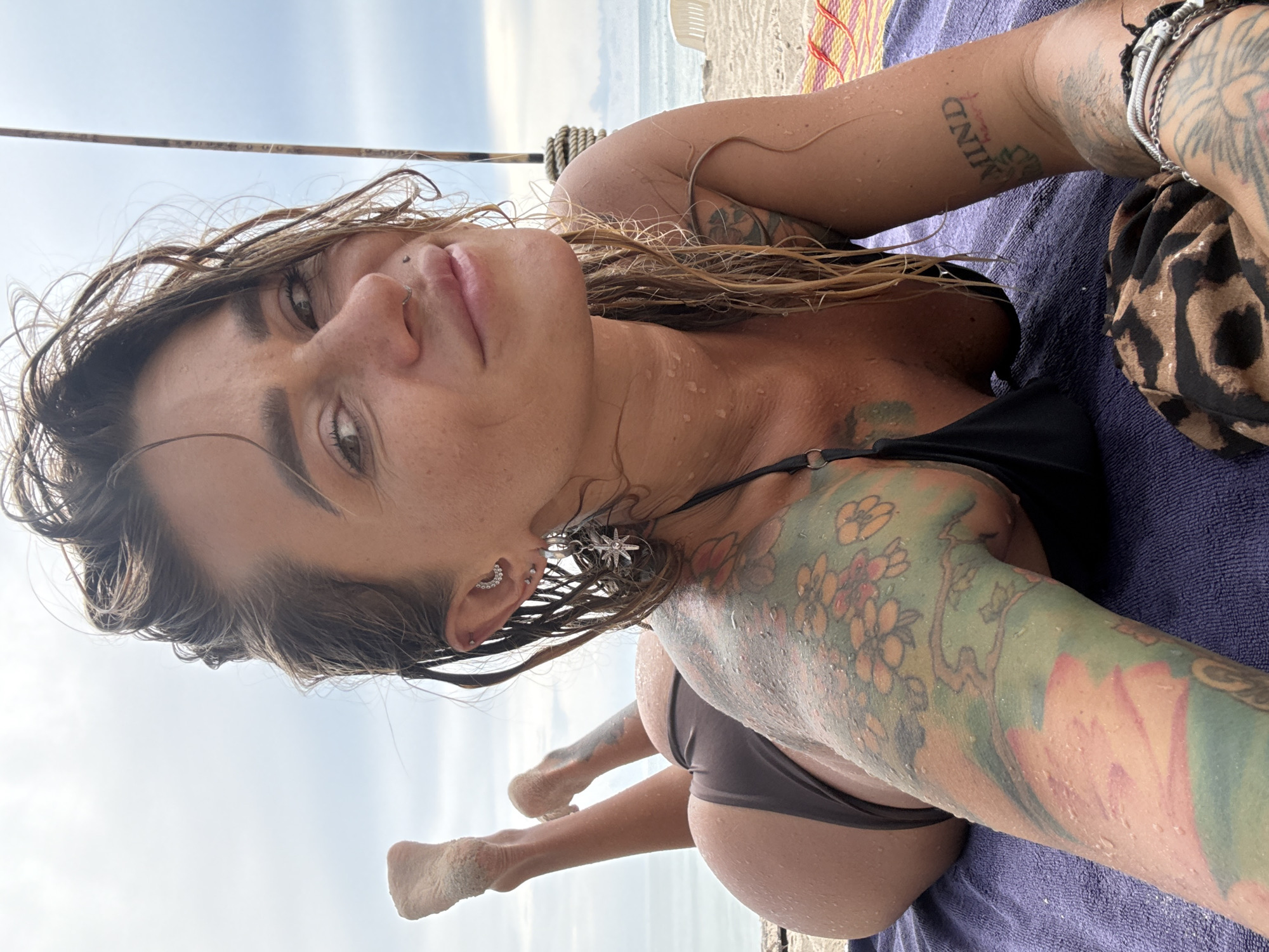 A woman with wet, brown hair and tattoos on her arms and chest lies face down on a purple towel on a sandy beach. She wears a dark bikini. Her feet are propped up, showing sand on her soles. The background shows a cloudy sky and the ocean. The scene appears to be in Choeng Thale, Thailand.