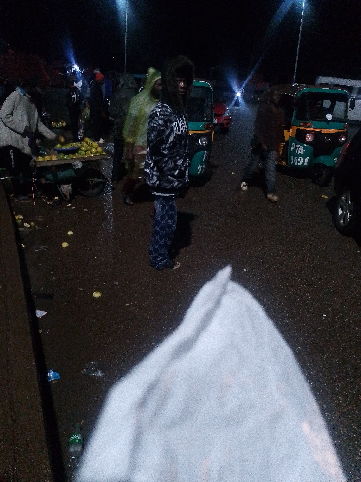 A night scene at a marketplace in Bukuru, Nigeria, shows a variety of people and vehicles. The ground is wet and appears to be asphalt, with scattered fruit, possibly oranges, and a discarded plastic bottle of Sprite. In the foreground, a person wearing patterned pants and a hooded jacket is the focus. Behind them, a bright light illuminates the scene from above. Several people are gathered around market stalls, with a fruit vendor visible in the mid-ground. Various modes of transportation are present, including a green and yellow auto-rickshaw with the license plate "PTA 9491" and another similar vehicle. Another auto-rickshaw is visible with the number "PT 77" on its side. A red vehicle is parked in the background. The overall atmosphere suggests a busy night market, likely operating after dark.