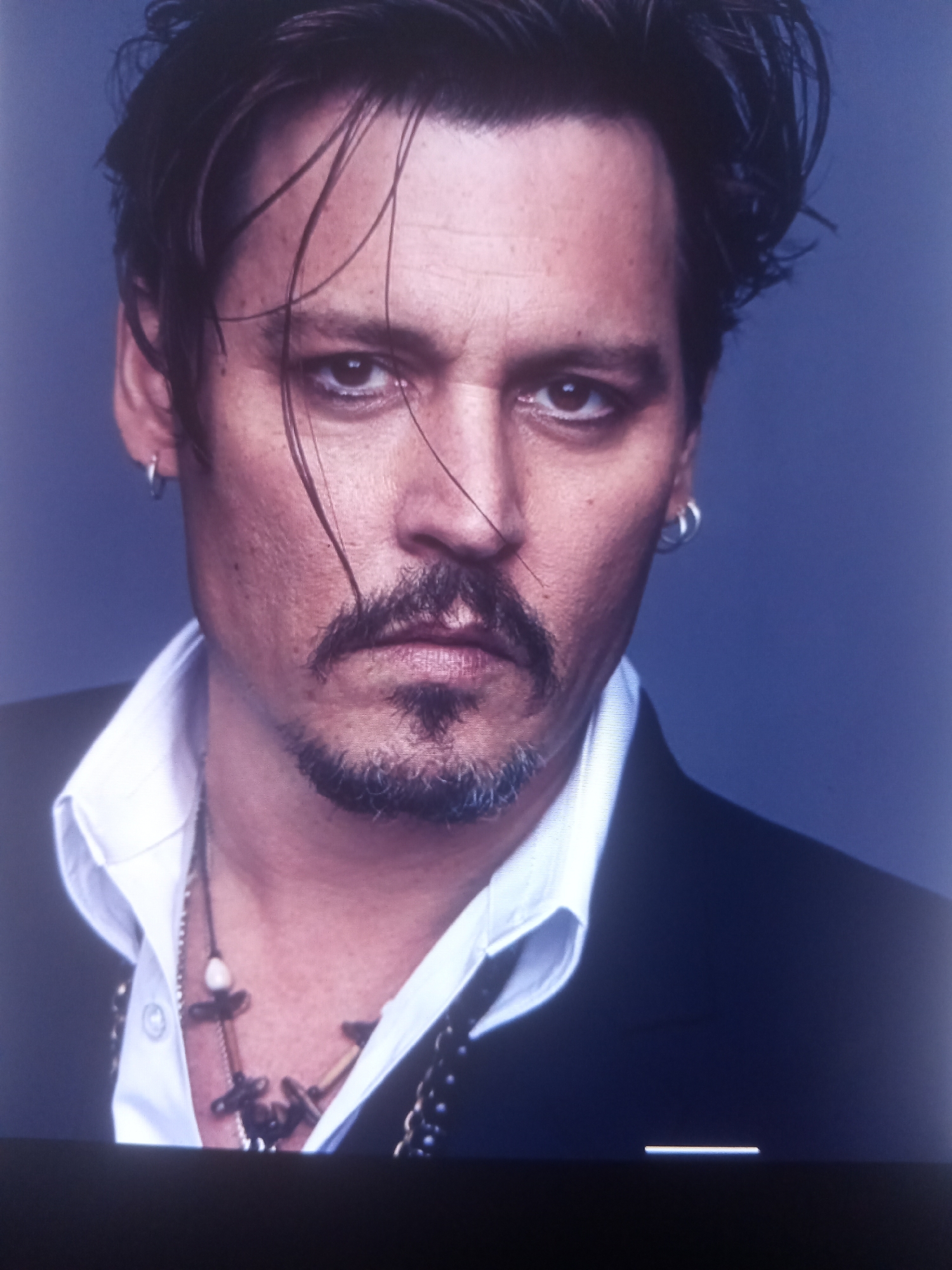 A medium shot shows a man with dark hair, styled with strands falling across his face. He is wearing a white collared shirt and a black jacket. He has hoop earrings in both ears and a necklace with several dark pendants and a white bead. The background is a solid, dark blue.