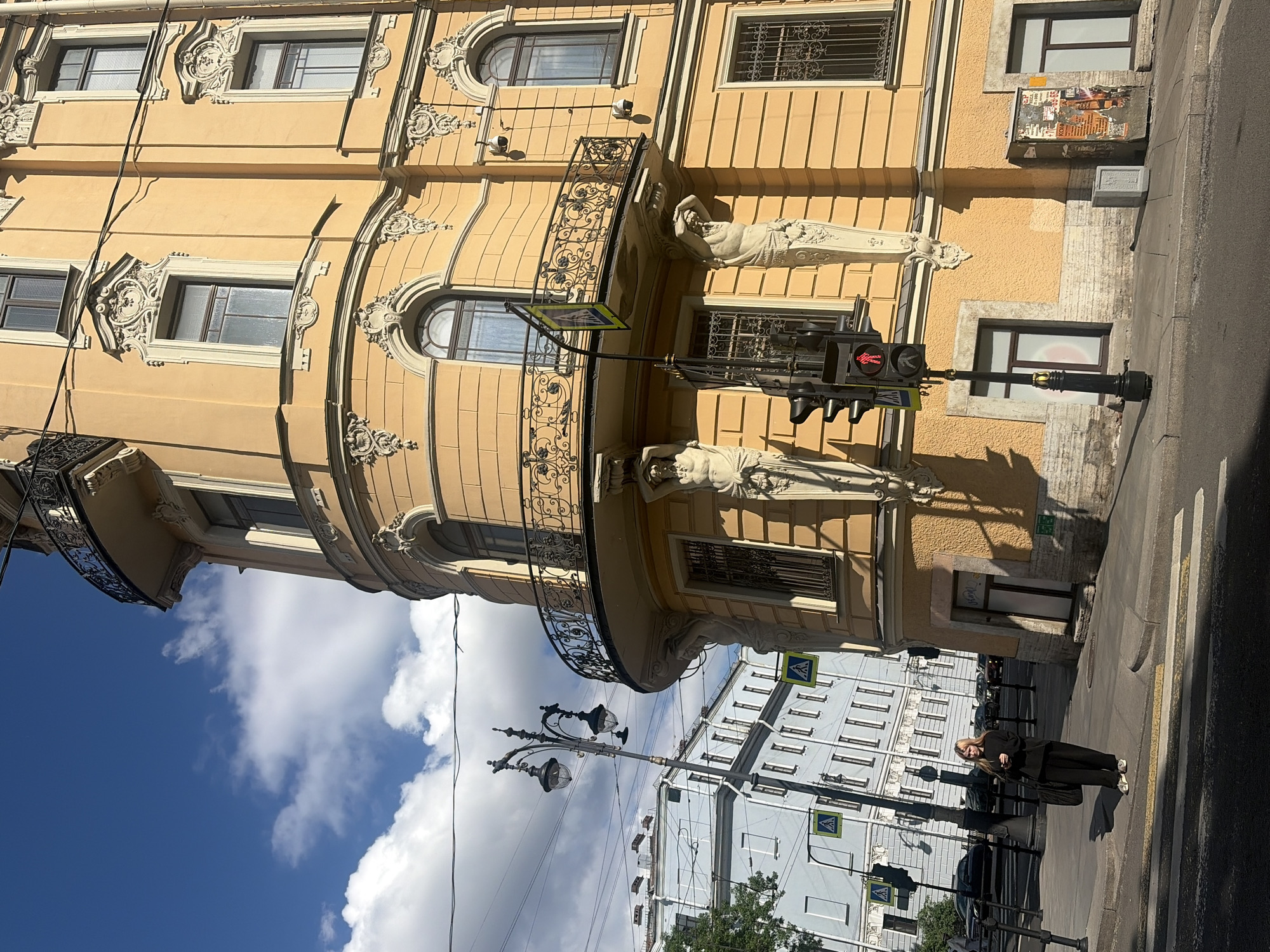 This is a street-level, eye-level, medium shot of an ornate, yellow building on a sunny day in Saint Petersburg, Russia. The building features elaborate architectural details including balconies with decorative metal railings, sculpted figures supporting balconies, and decorative window surrounds. The building's facade is a bright yellow color. On the sidewalk in front of the building, a woman wearing black pants and a black sweater stands looking at her phone. She is carrying a large black bag over her shoulder. A traffic light with a red walking figure illuminated is visible on a pole in the foreground. Across the street, another building with a light blue facade is visible. The sky is blue with scattered white clouds. Overhead wires are visible strung between poles.