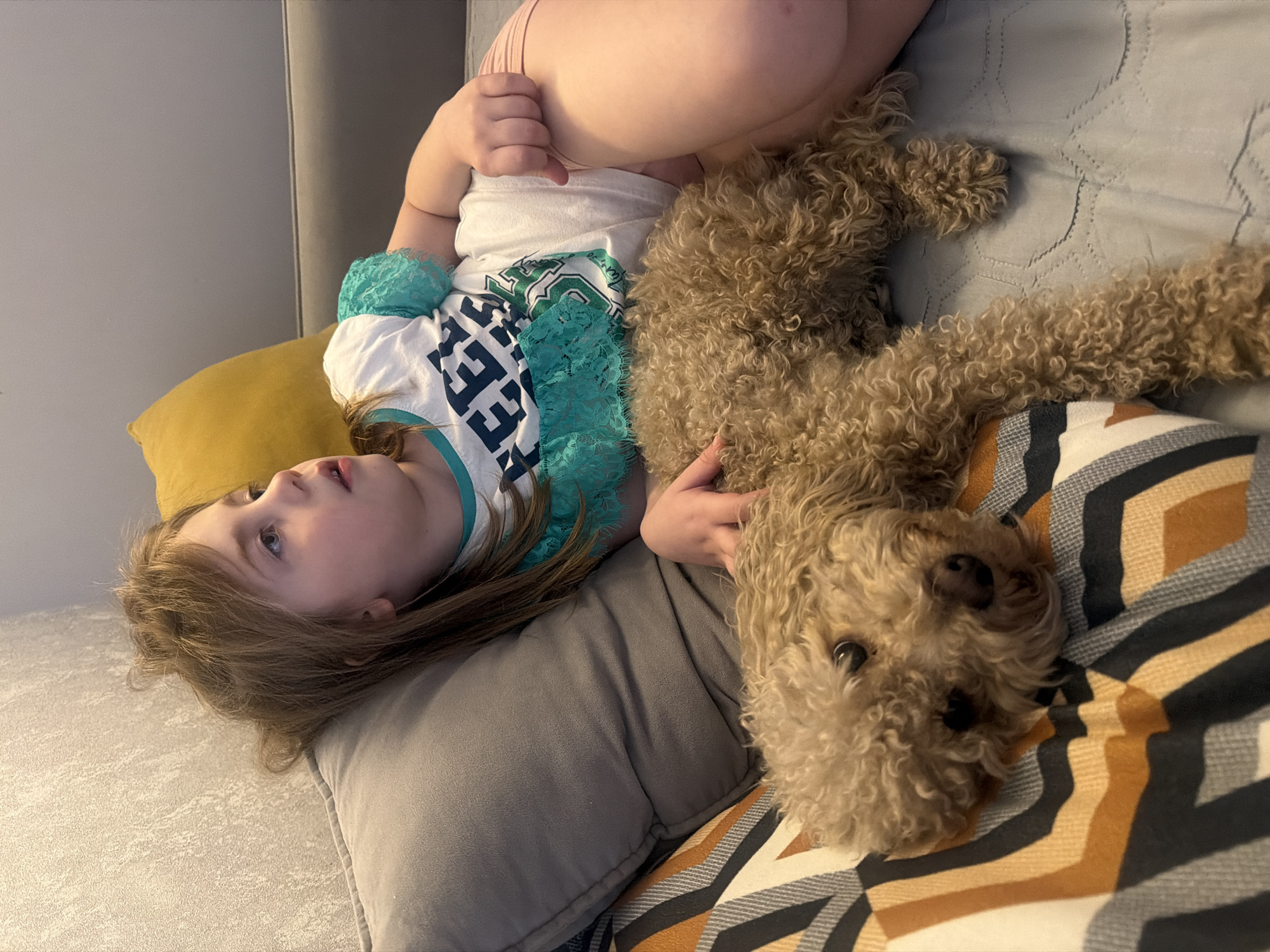 A child and a poodle are lying on a couch. The child is wearing a white t-shirt with a green design and blue lace trim. The poodle is light brown with curly hair. The couch has a yellow pillow, a gray pillow, and a patterned throw. The location is not specified and cannot be determined from the image.