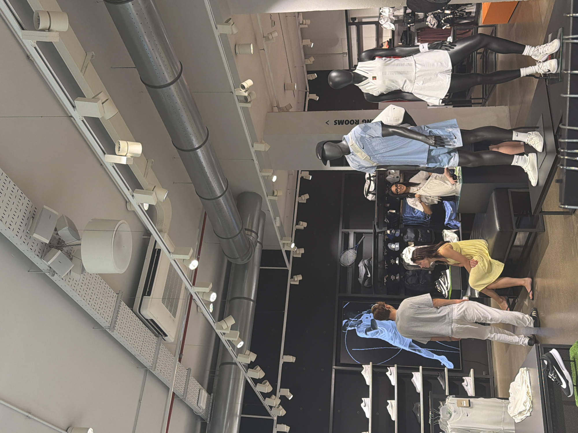 The image depicts the interior of a Nike store in Milan, Italy. The store features high ceilings with visible ventilation ducts and lighting fixtures. On display are various Nike products, including apparel and footwear, showcased on shelves, racks, and mannequins. Several mannequins are dressed in athletic wear, some arranged as a display. In the foreground, a black mannequin is dressed in a white tennis outfit, and another is in a light blue polo shirt and shorts. In the midground, a man and a woman are interacting. The man, wearing a grey t-shirt and white pants, is standing, while the woman, in a yellow dress, is seated on a bench. A female employee is visible behind a counter in the background, holding apparel. The store's aesthetic is modern and clean, with a focus on product presentation.