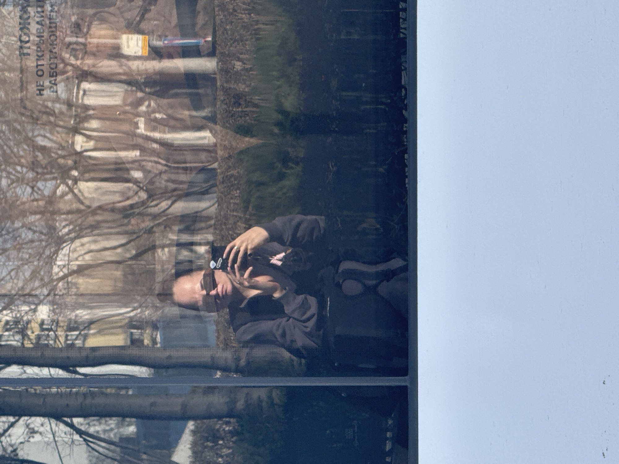 The image shows a reflection in a window. The reflection includes a person holding a camera and taking a photo. The reflection also shows trees and buildings. A sign with the words "НЕ ОТКРЫВАЙТЕ РАБОТАЮЩИЕМ" is visible on the window. The location is Moscow, Russia.
