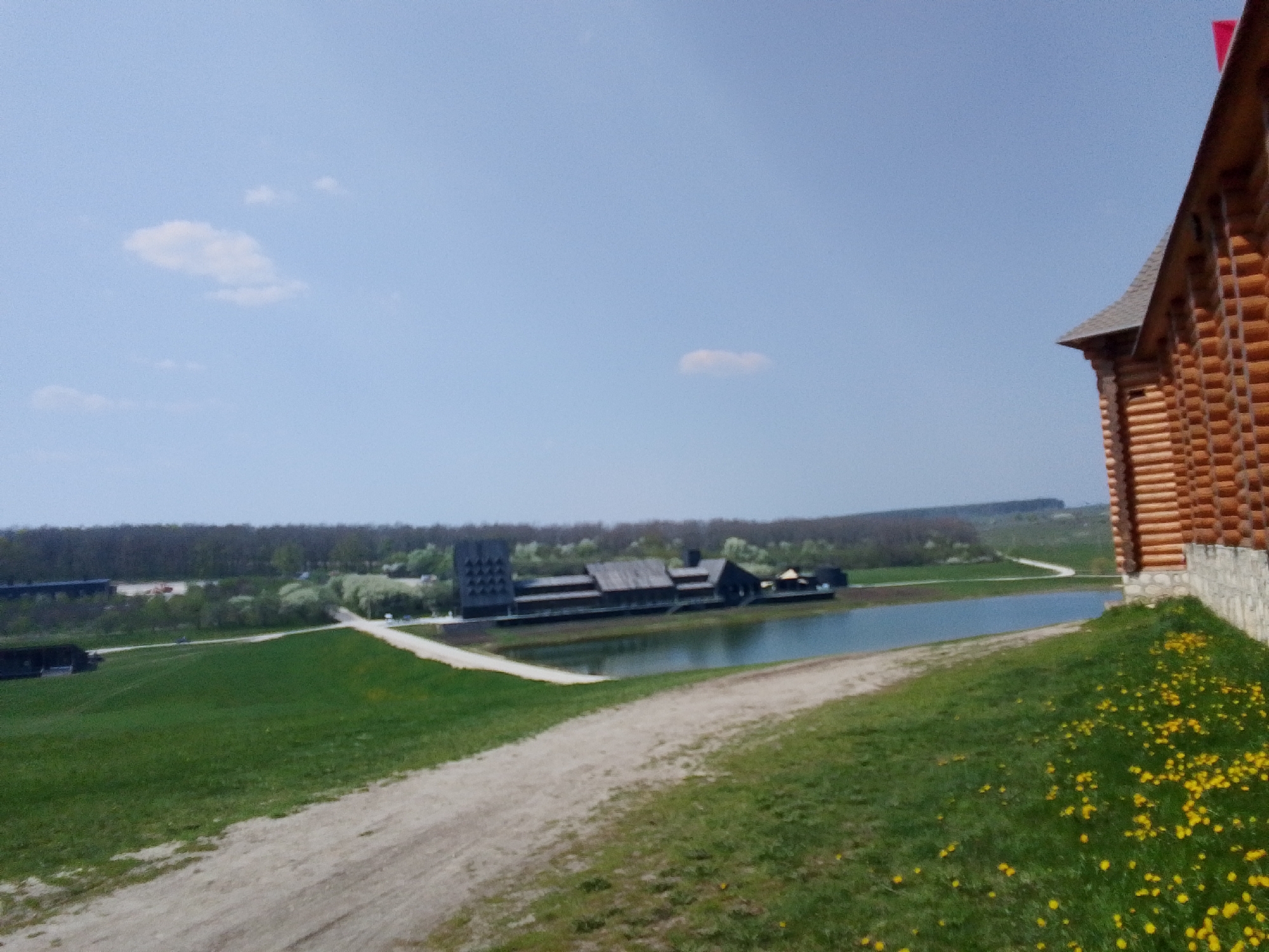 The image shows a landscape in Каменка, Russia. A dirt path leads from the foreground towards a pond. On the far side of the pond is a long building with a distinctive tower-like structure. The building appears to be constructed with dark wood and is partially reflected in the water. Trees are visible along the horizon, forming a backdrop to the scene. To the right in the foreground, there is a portion of a log building. The grass is green with scattered yellow flowers. The sky is a clear blue with a few small clouds.