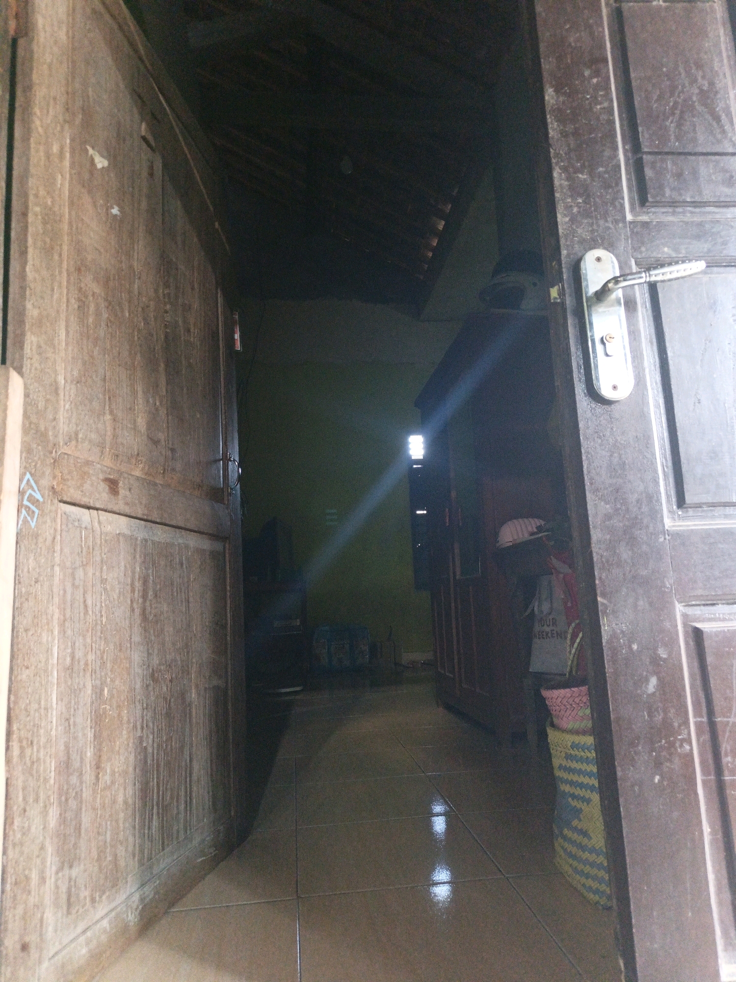 The image depicts an interior view looking into a dimly lit room from a doorway. On the left, a large wooden door, likely part of a cabinet or closet, is ajar, revealing a weathered wooden surface with some faint blue markings. The door on the right is also wooden and open, featuring a metallic door handle and lock mechanism. The floor is tiled with a brown, glossy finish, reflecting light.
The room beyond the doorway is dark, with a strong diagonal light beam originating from a source to the left and extending across the scene, illuminating dust particles in the air. In the background, a dark wooden cabinet or shelving unit is visible, and on top of it, a pink and yellow woven basket sits atop a larger woven cylindrical container with a blue and yellow patterned design. A white hat with a red ribbon is perched on the side of the cabinet. Further into the room, there are hints of other objects and structures, including what appears to be a television set or similar electronic device. The ceiling is visible in the upper part of the image, constructed with dark wooden beams and a thatched or similar material. The scene appears to be in a residential setting, possibly a home or storage area. The location of Kamulyan, Indonesia, is mentioned as context for this scene.